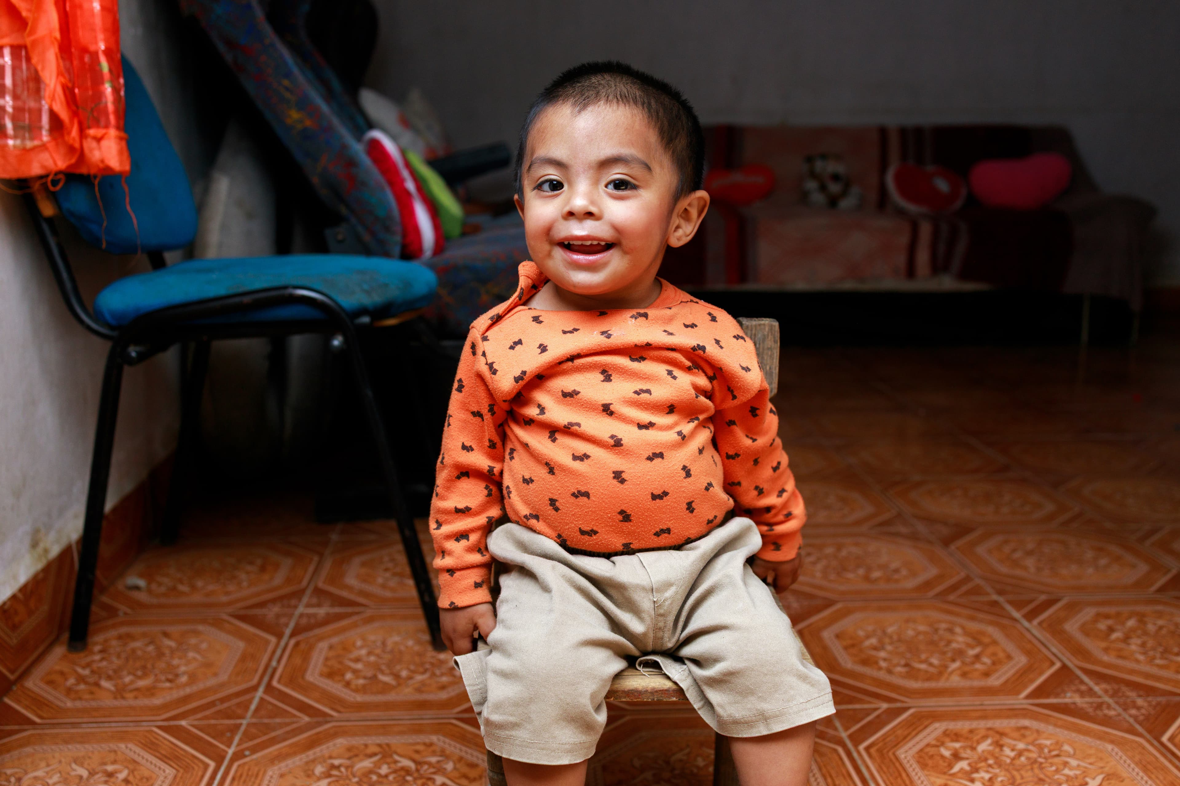 A toddler Mexican boy sits in a wooden chair and smiles for the camera while wearing a bright orange shirt.