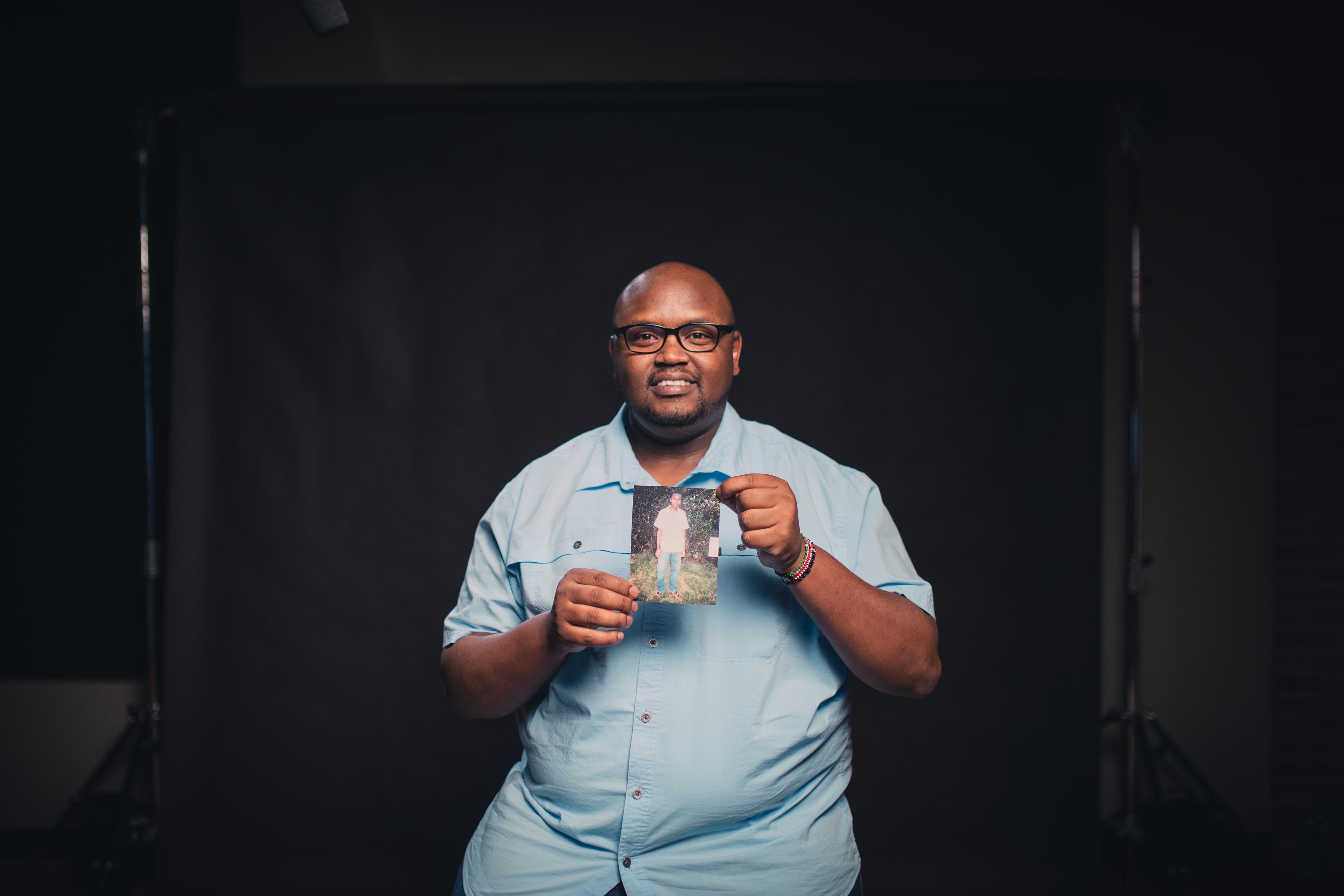 A man is sitting and smiling as he holds up a photo of himself as a child.