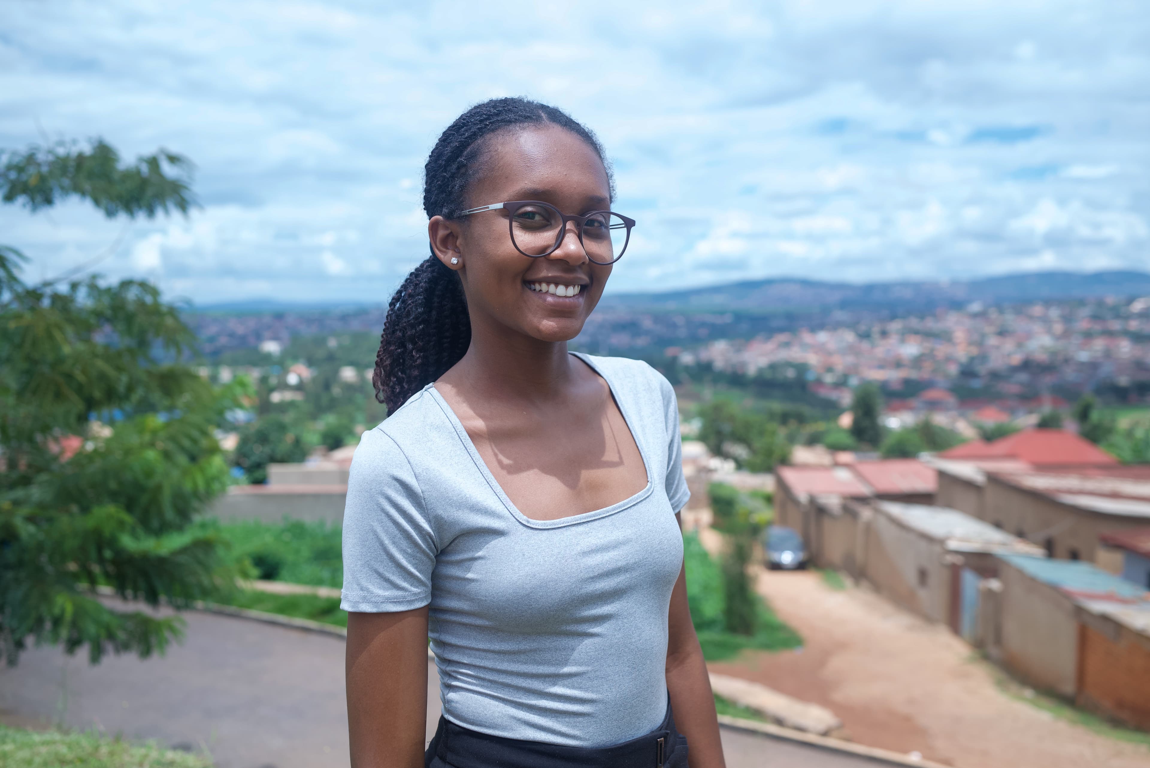 An older teenaged girl smiles at the camera while standing outdoors. She is on a hill overlooking houses, mountains and trees.