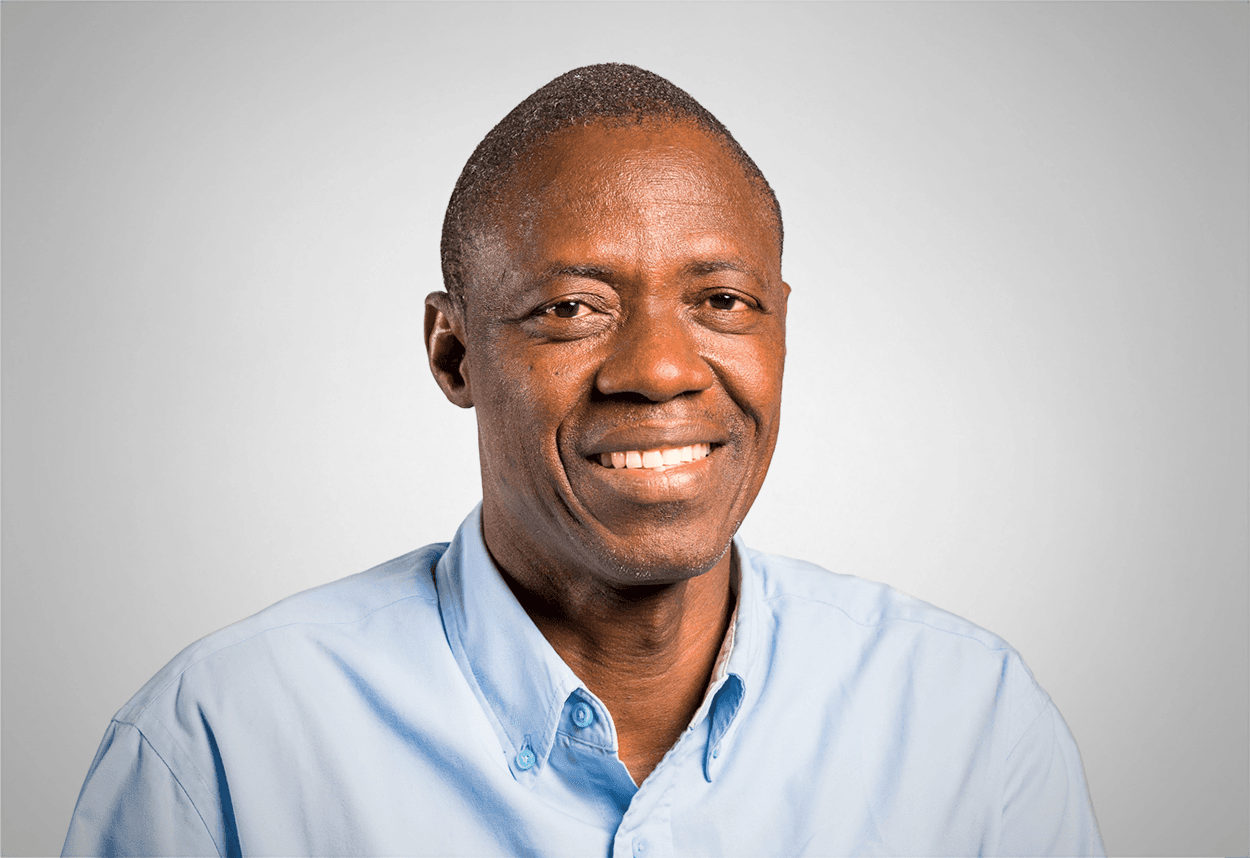 A man in a blue button up shirt is smiling, with a grey background behind him.