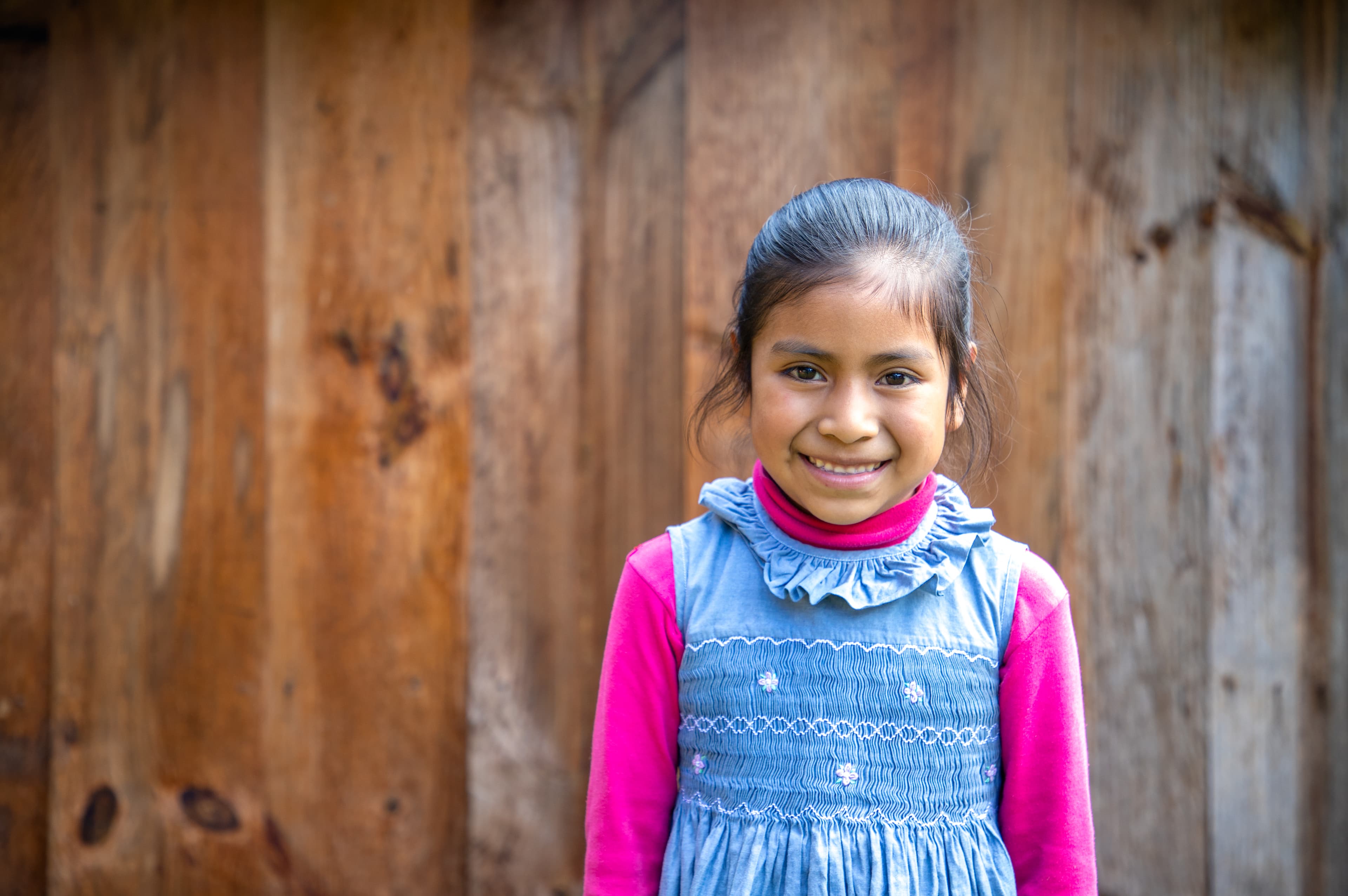 A young Mexican girl wearing a denim dress over a pink shirt smiles for the camera.