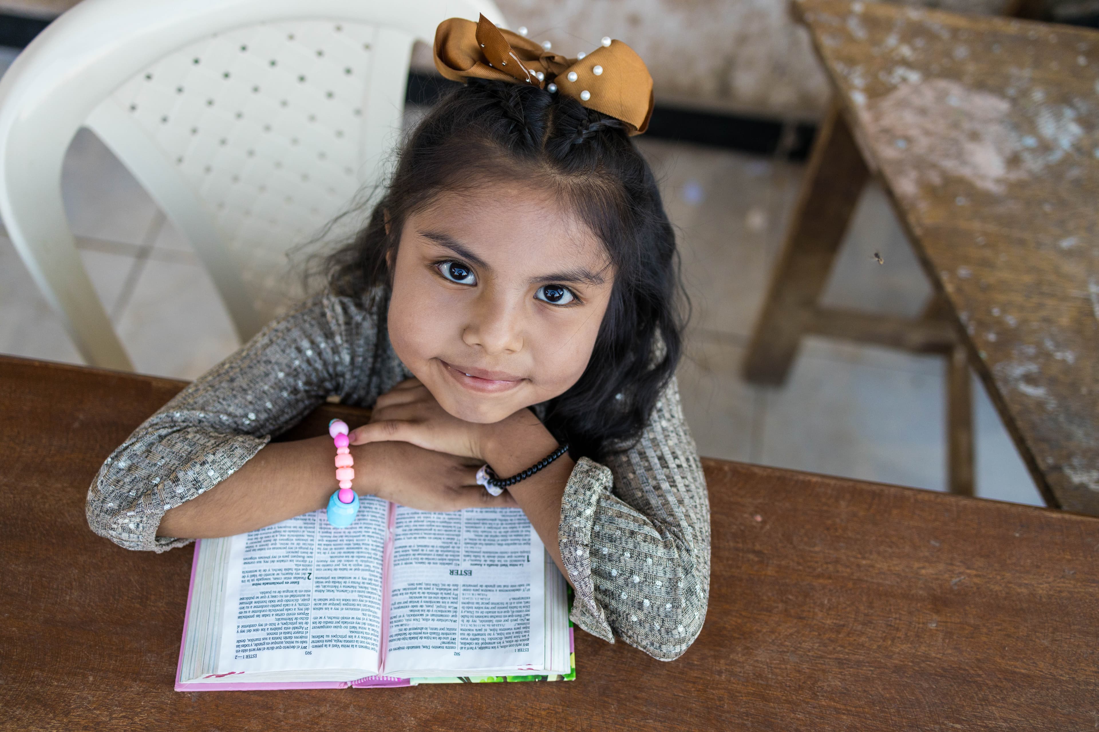 A young girl is sitting at a desk and resting her hands on a bible as she looks at the camera.