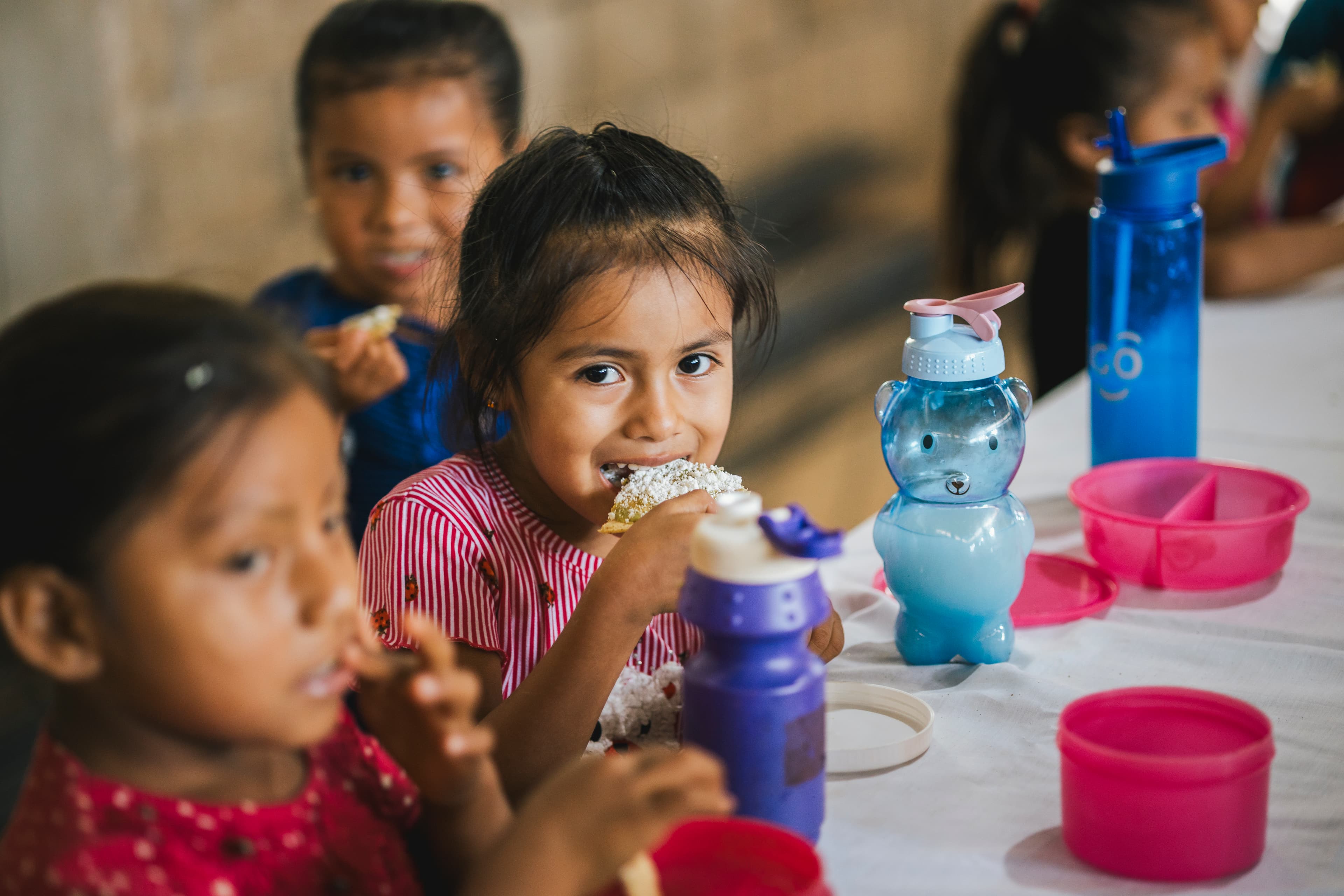 A young girl is sitting at a table and eating a piece of bread surrounded by classmates.