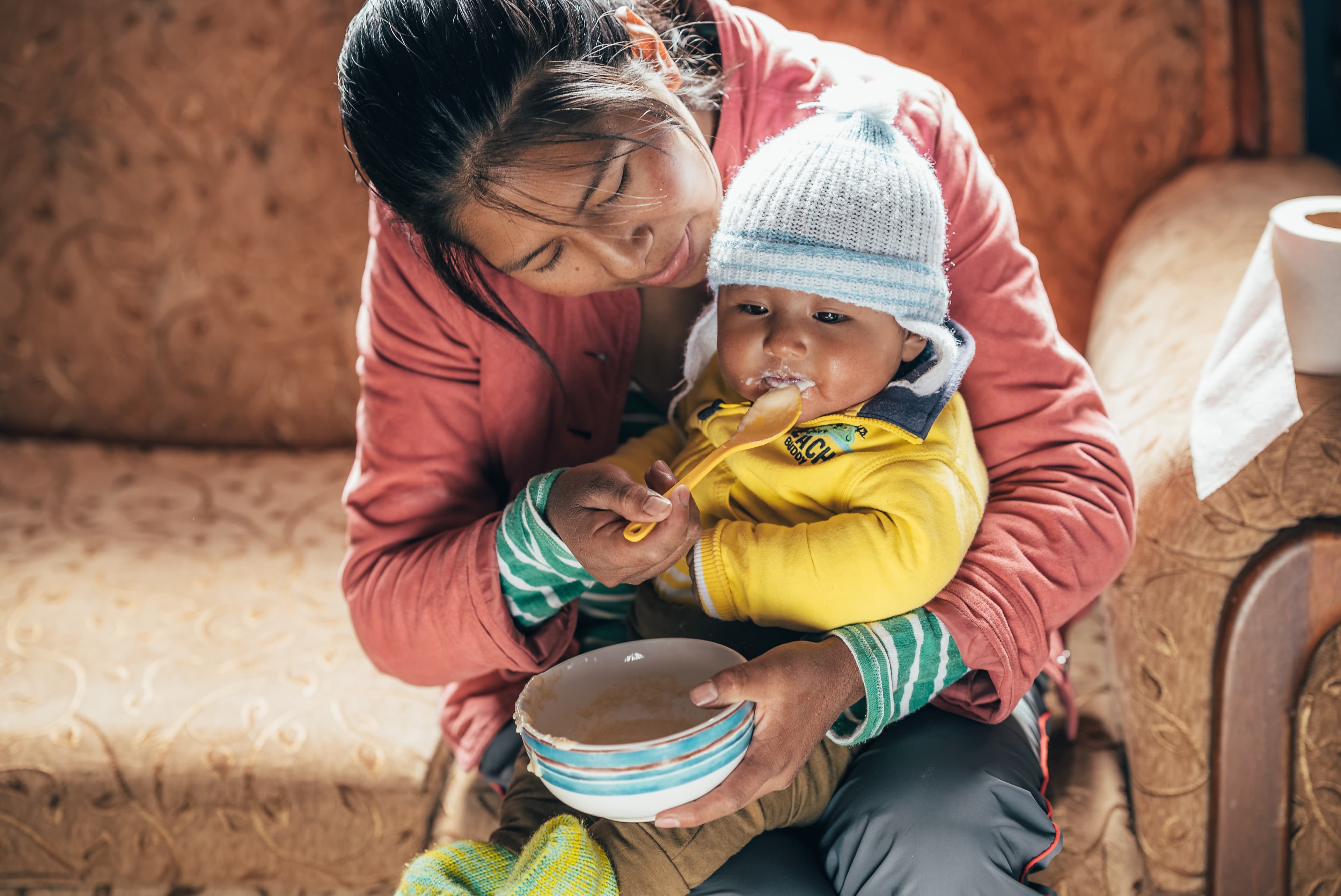 A mother, in a pink jacket, is feeding her baby boy, who is wearing a yellow jacket and white knit hat.