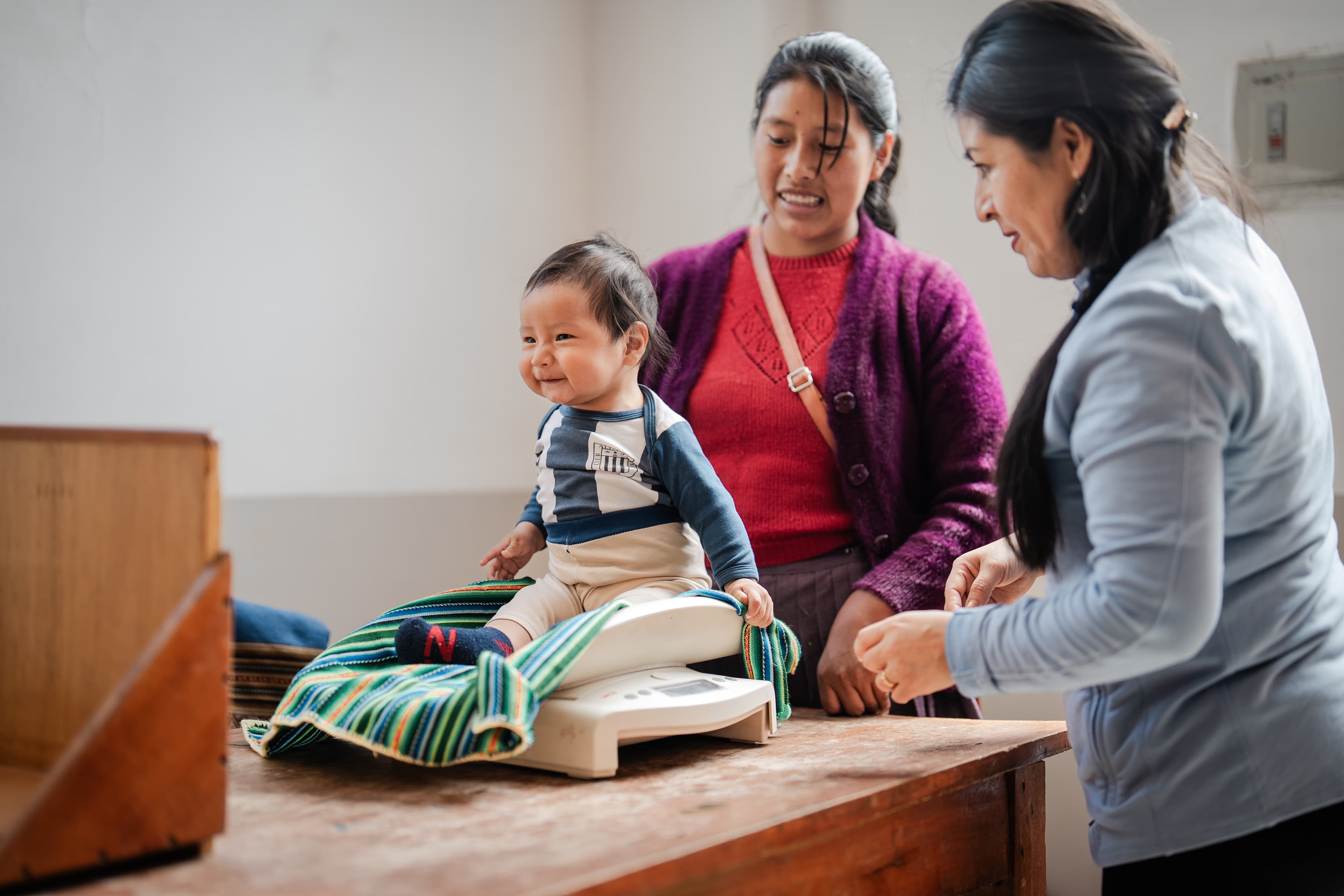 A mom stands behind her baby boy, who is smiling while being weighed on a scale on top of a wooden table. A woman helping them stands next to them.