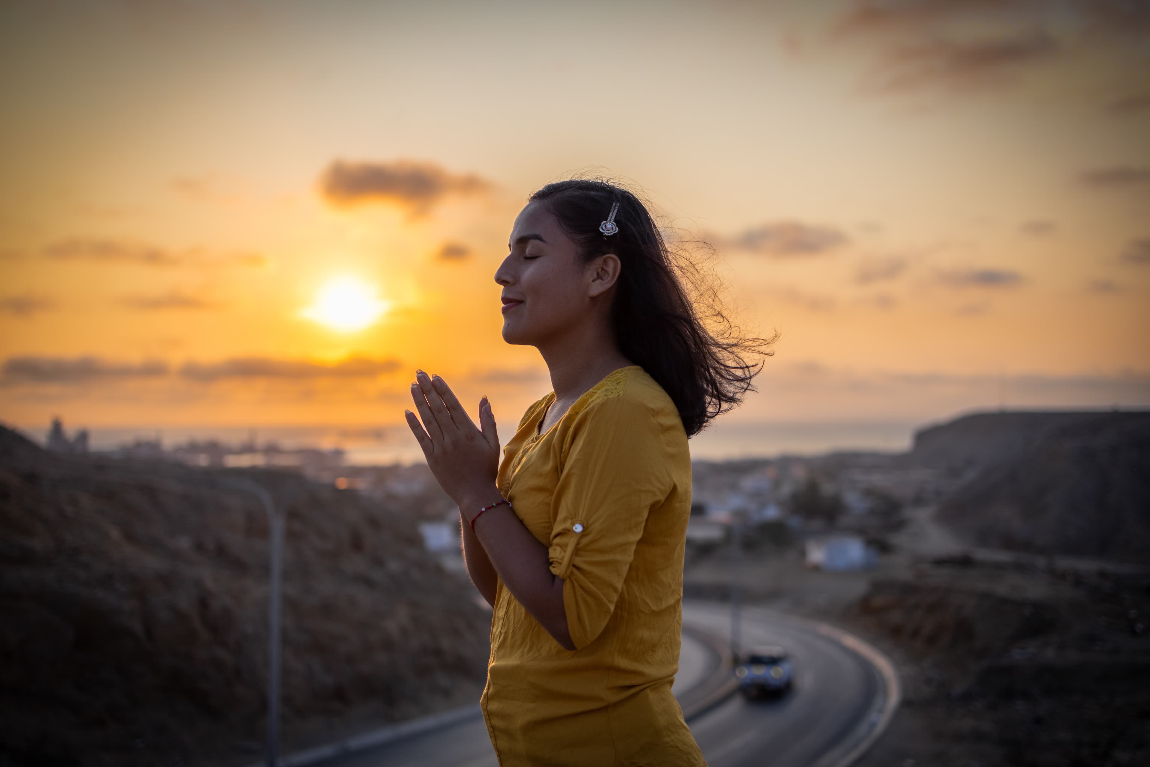 A teen girl clasps her hands together and prays in front of a bright sunset.