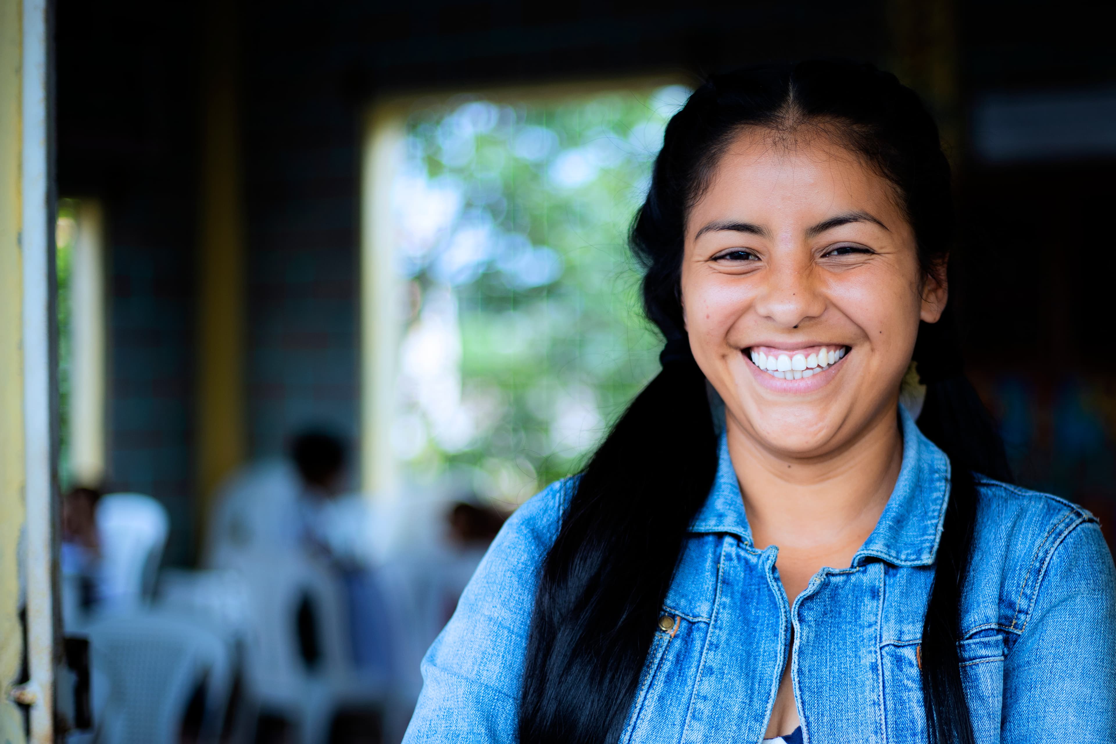 A young woman smiles at the camera. She is wearing a blue jean jacket and her hair in two ponytails.