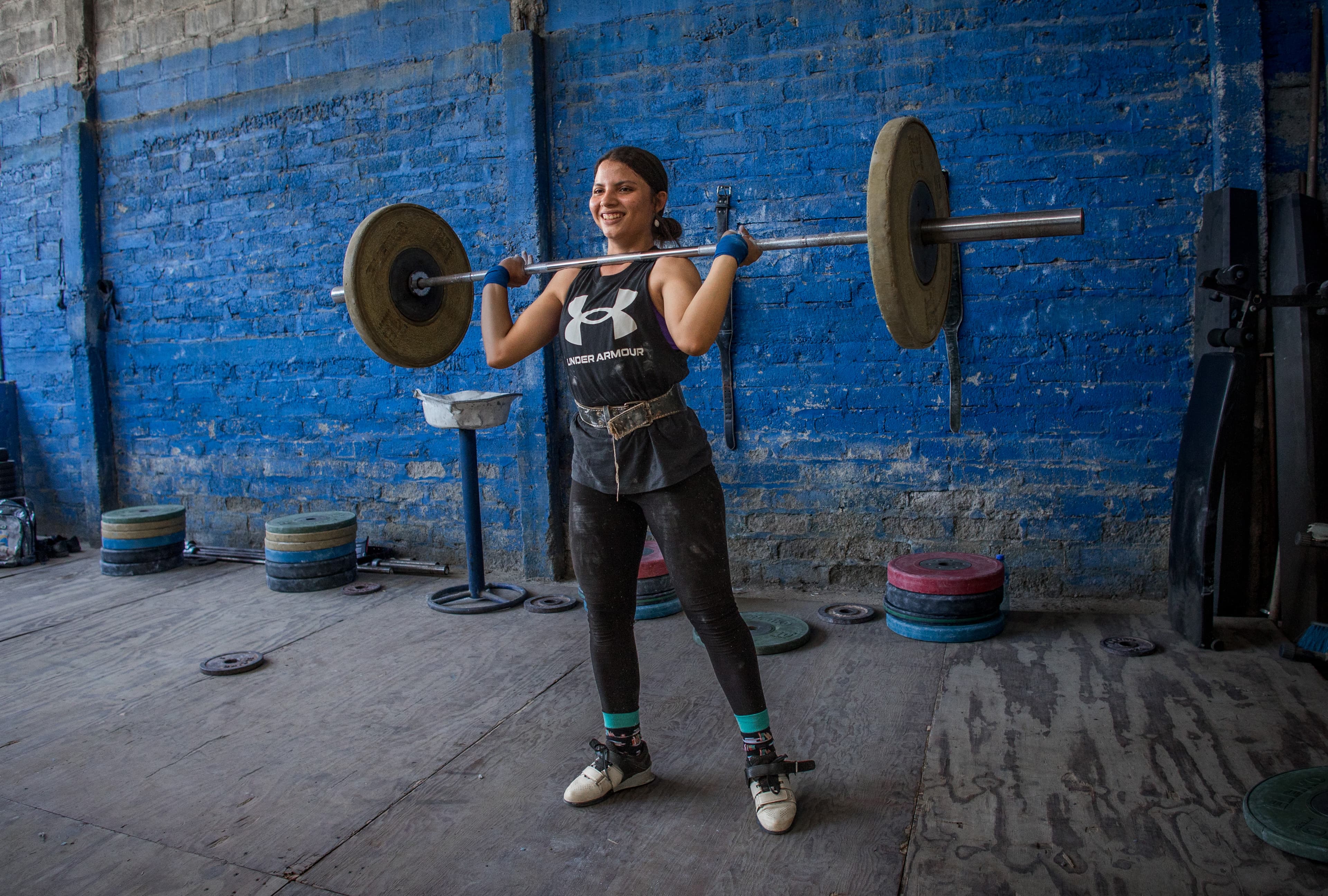 A girl lifts weights up to her shoulders while smiling.