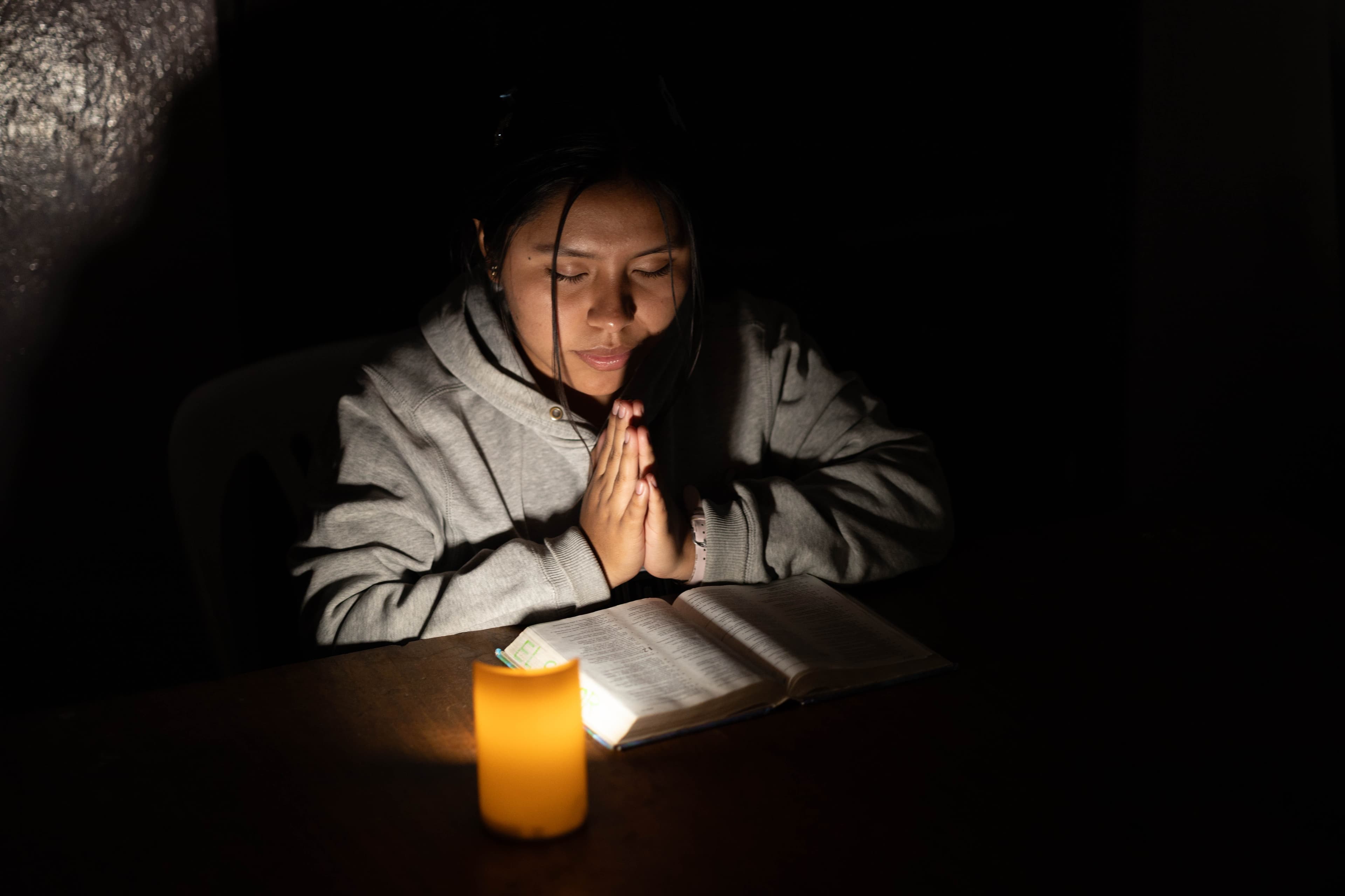 A teenage girl sits at a table in a dark room praying, illuminated by a candle, with her Bible open in front of her.