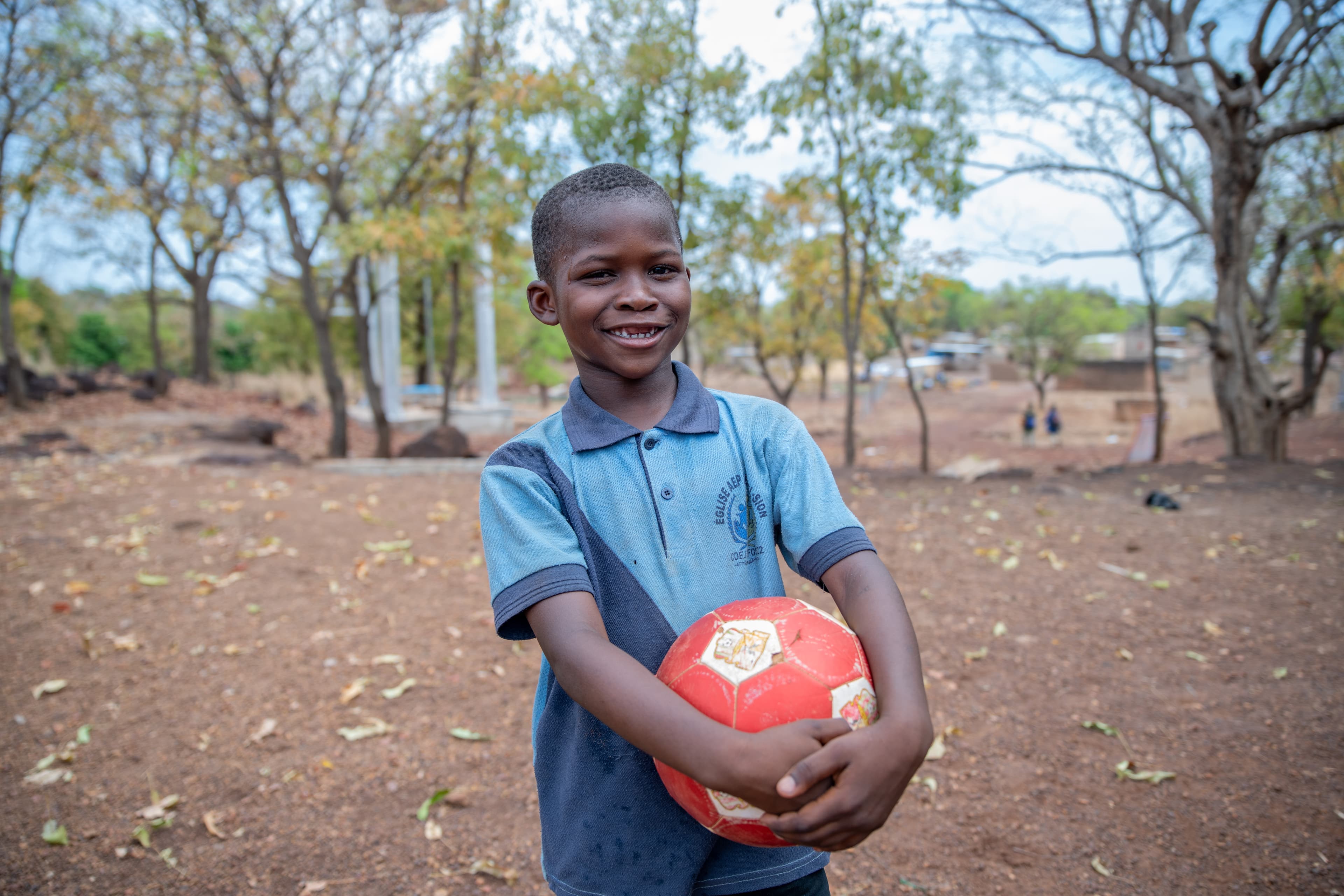 A young African boy wearing a blue polo holds a red soccer ball.