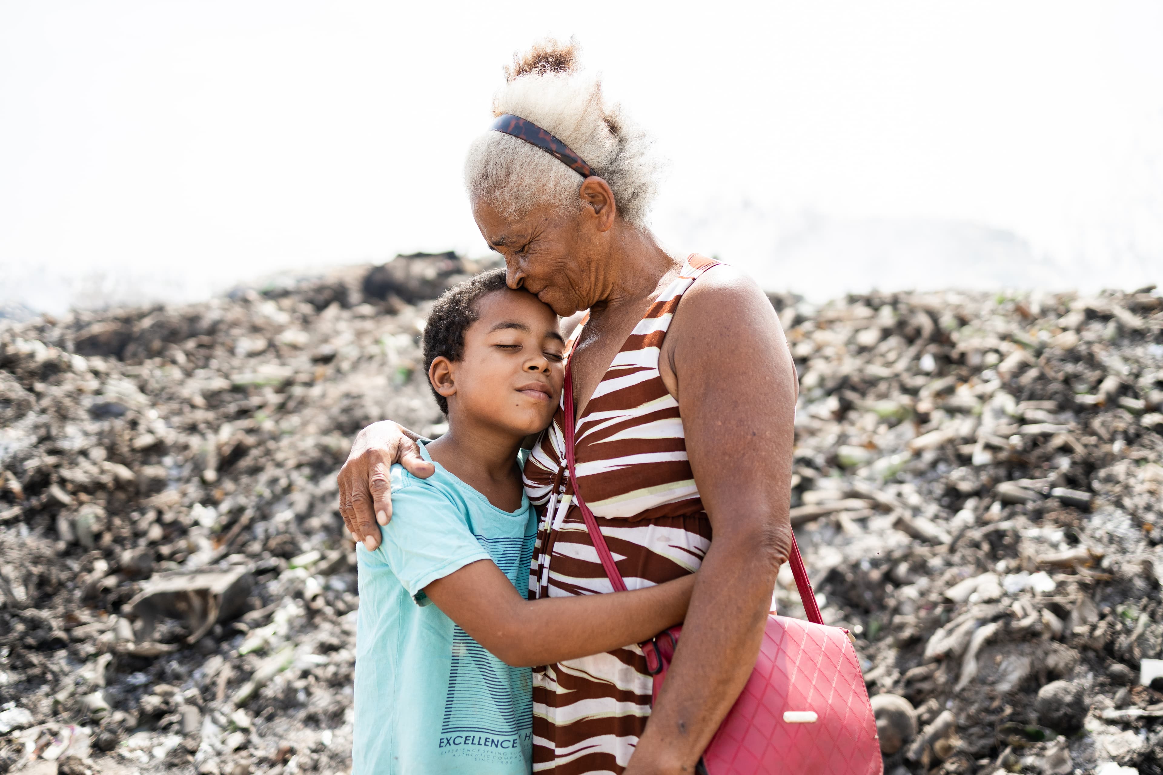 An elderly woman hugs a young boy in front of a garbage pile.