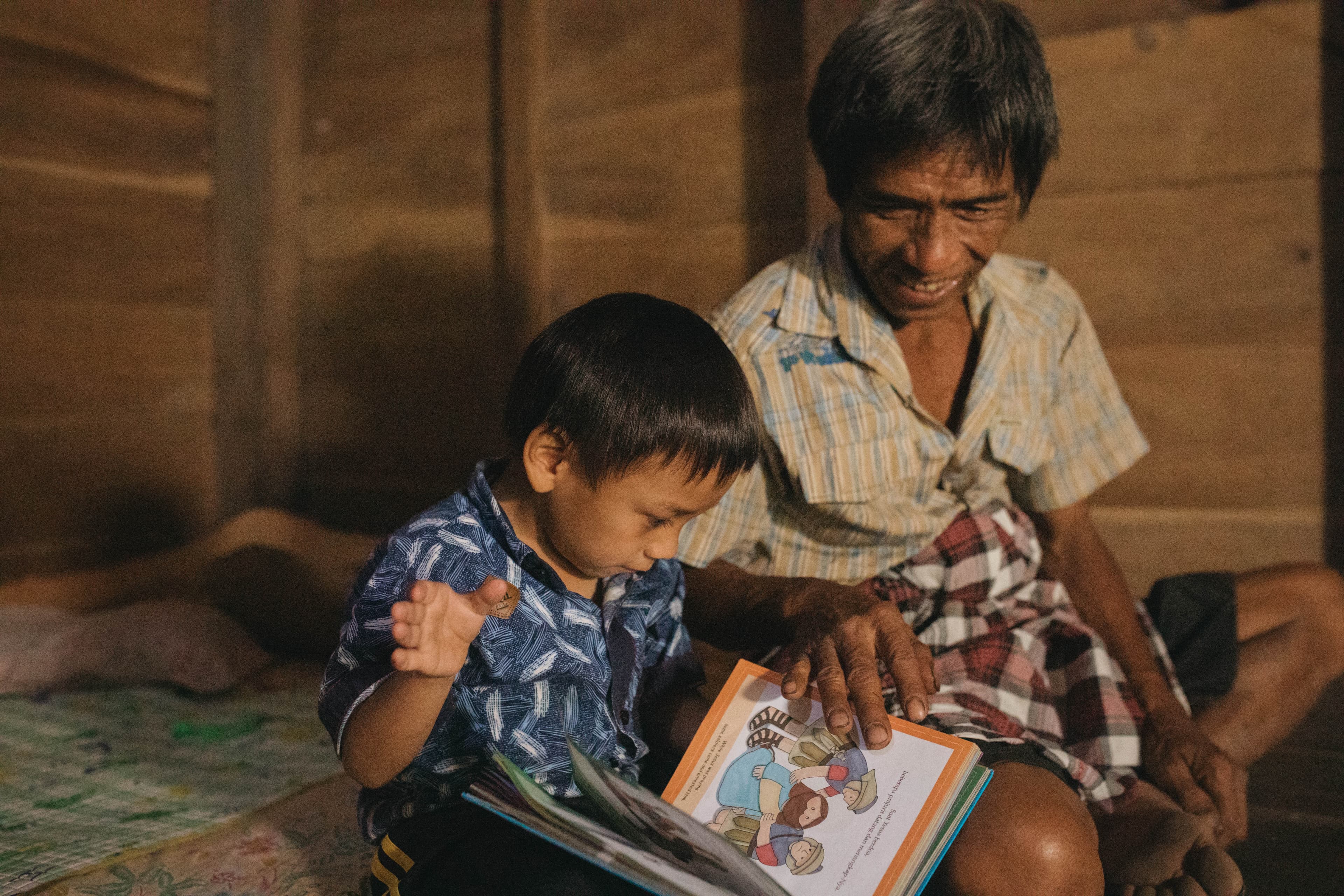 A young boy sits next to an older man as they look through an illustrated Bible.