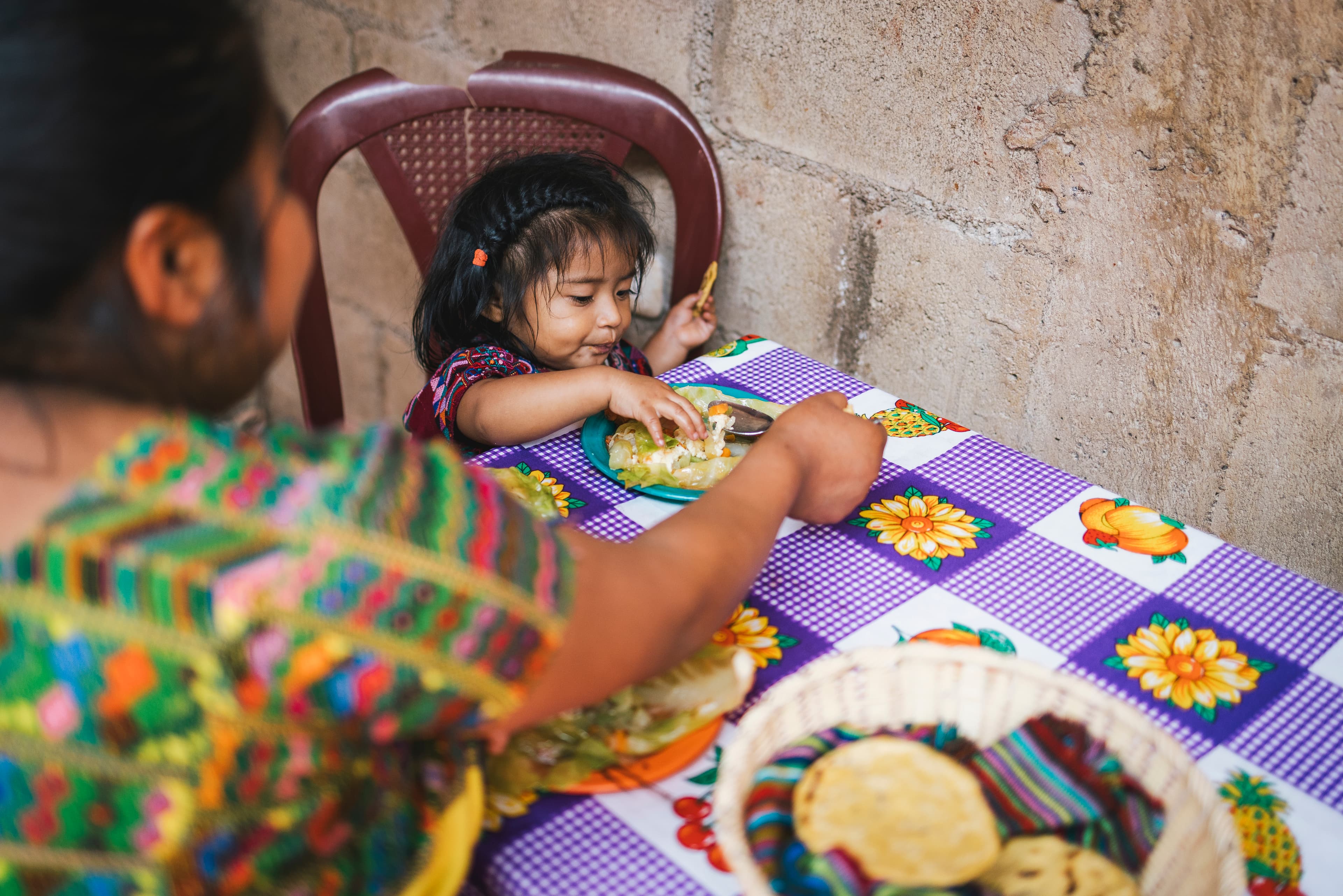 A little girl is sitting at a table eating food with her mother.