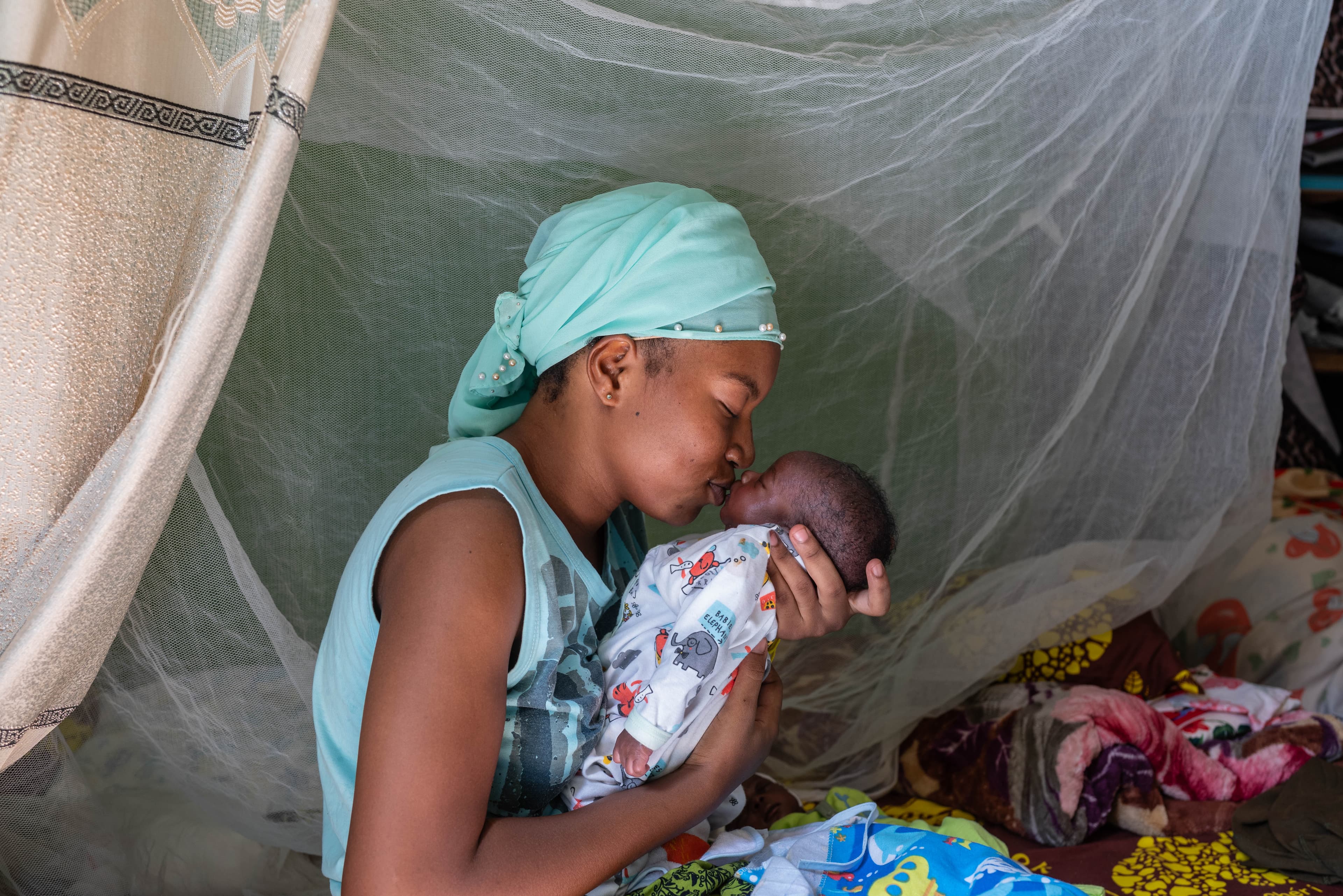 A mom holds and kisses her infant child while surrounded by a mosquito net.