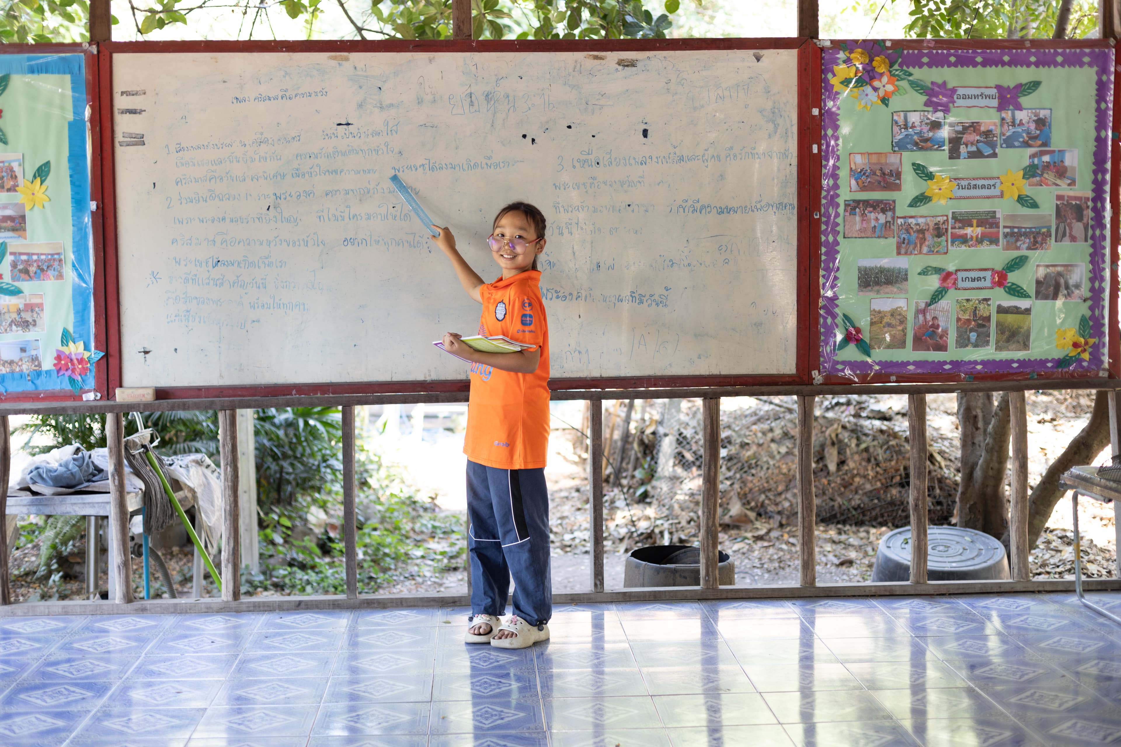 A young girl wearing a bright orange shirt holds a book and points to a whiteboard while smiling.