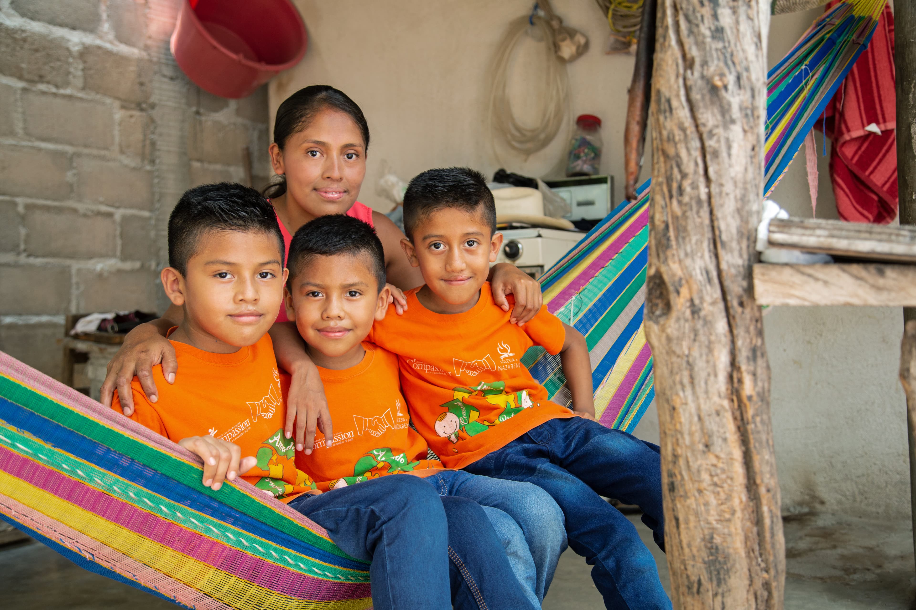 Three young boys are sitting in a hammock outside their home with their mother behind them.