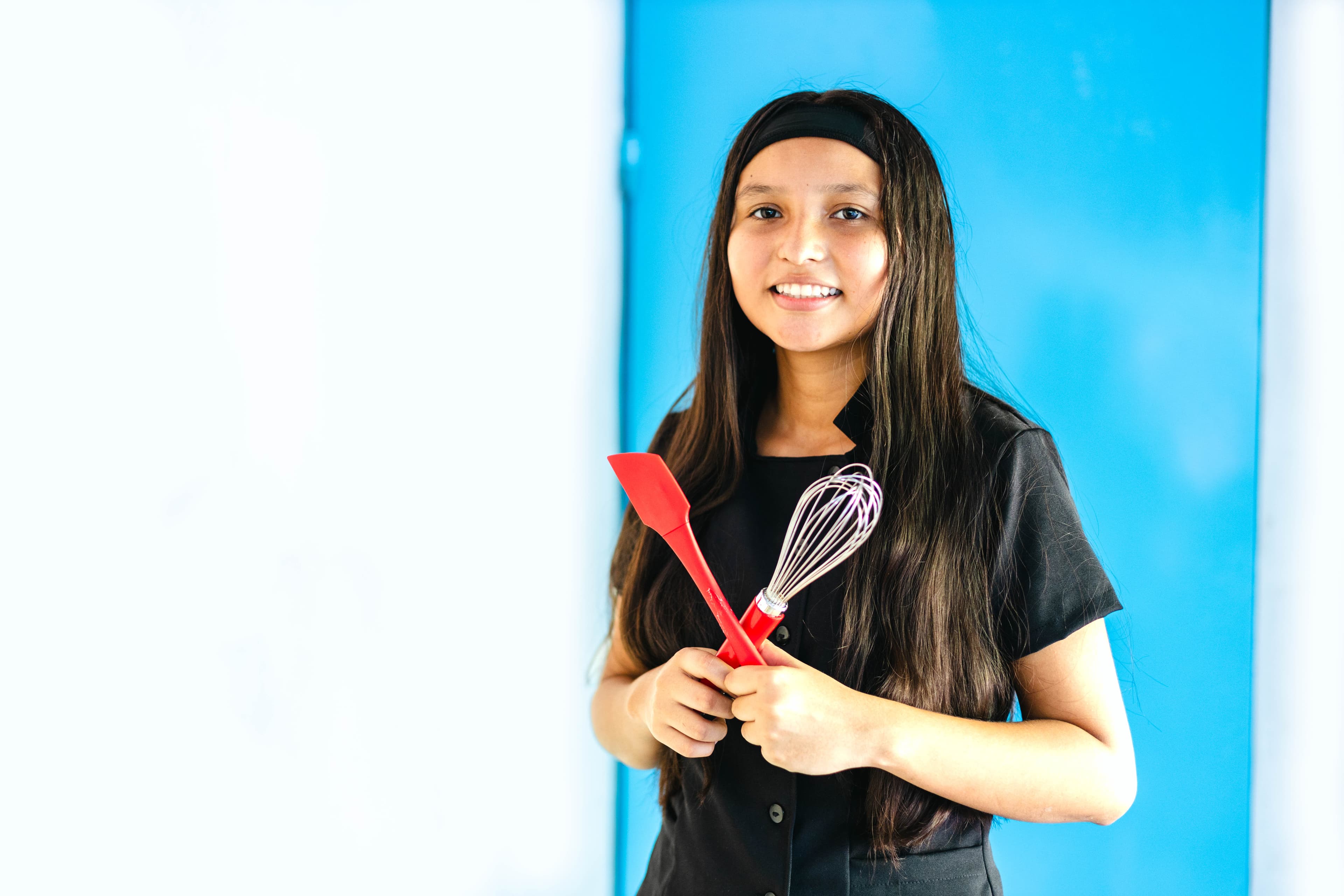 A young Salvadoran girl holds a whisk and an icing spatula while smiling in front of a blue wall.