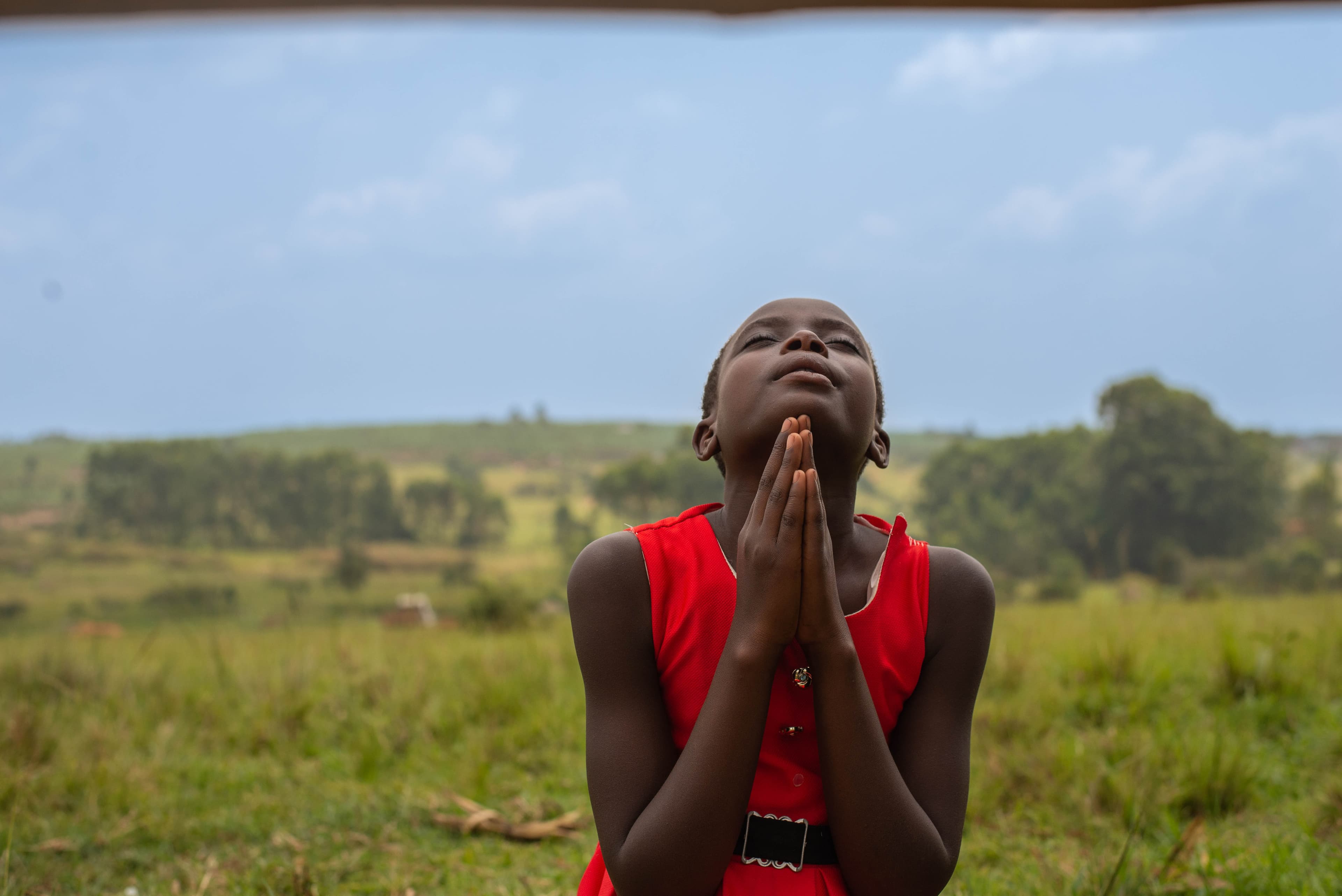A girl wearing a red dress with a black belt is sitting outside with her hands folded in prayer. Her head is tilted upward.