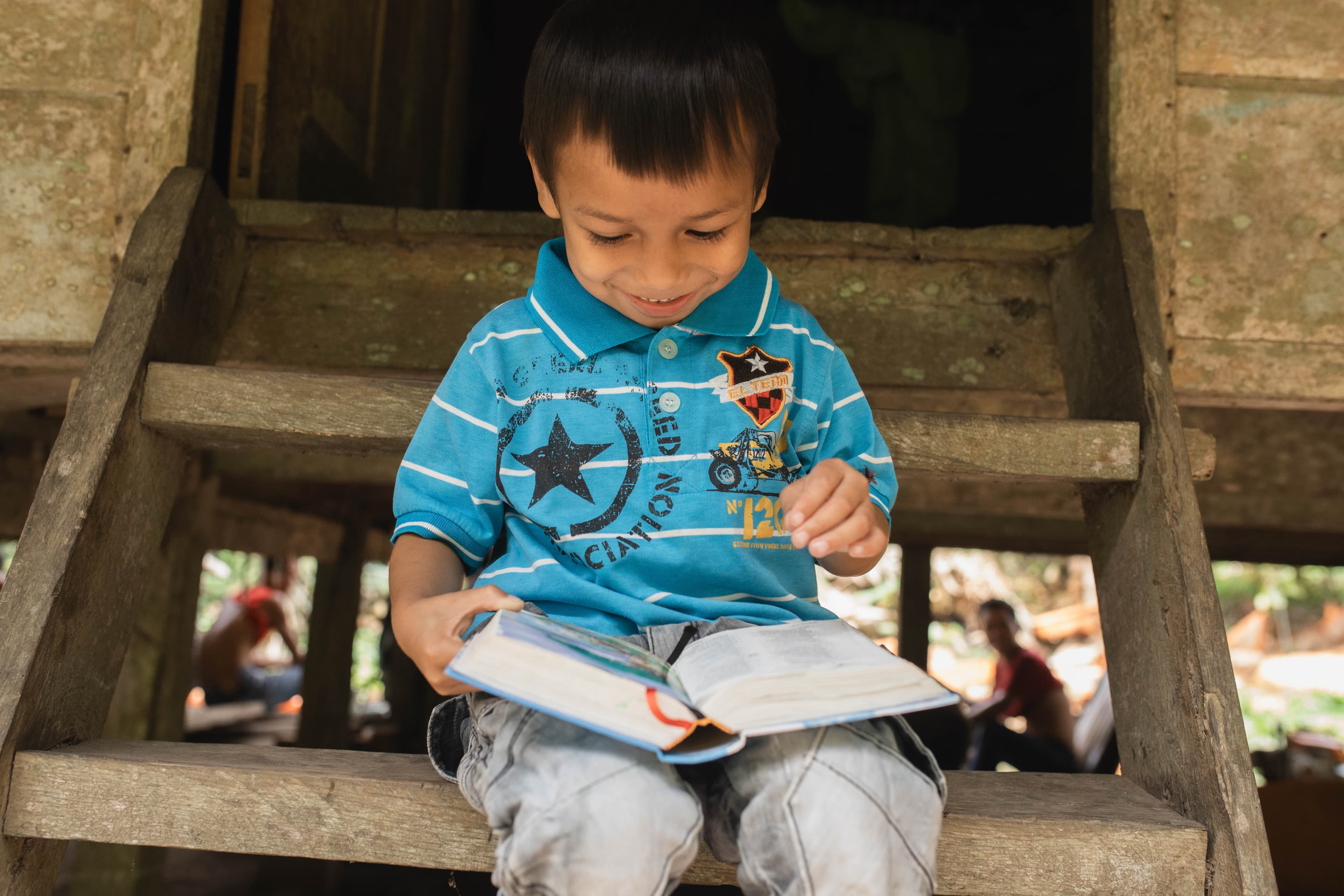 A little boy sits outside on a staircase while reading a Bible and smiling.