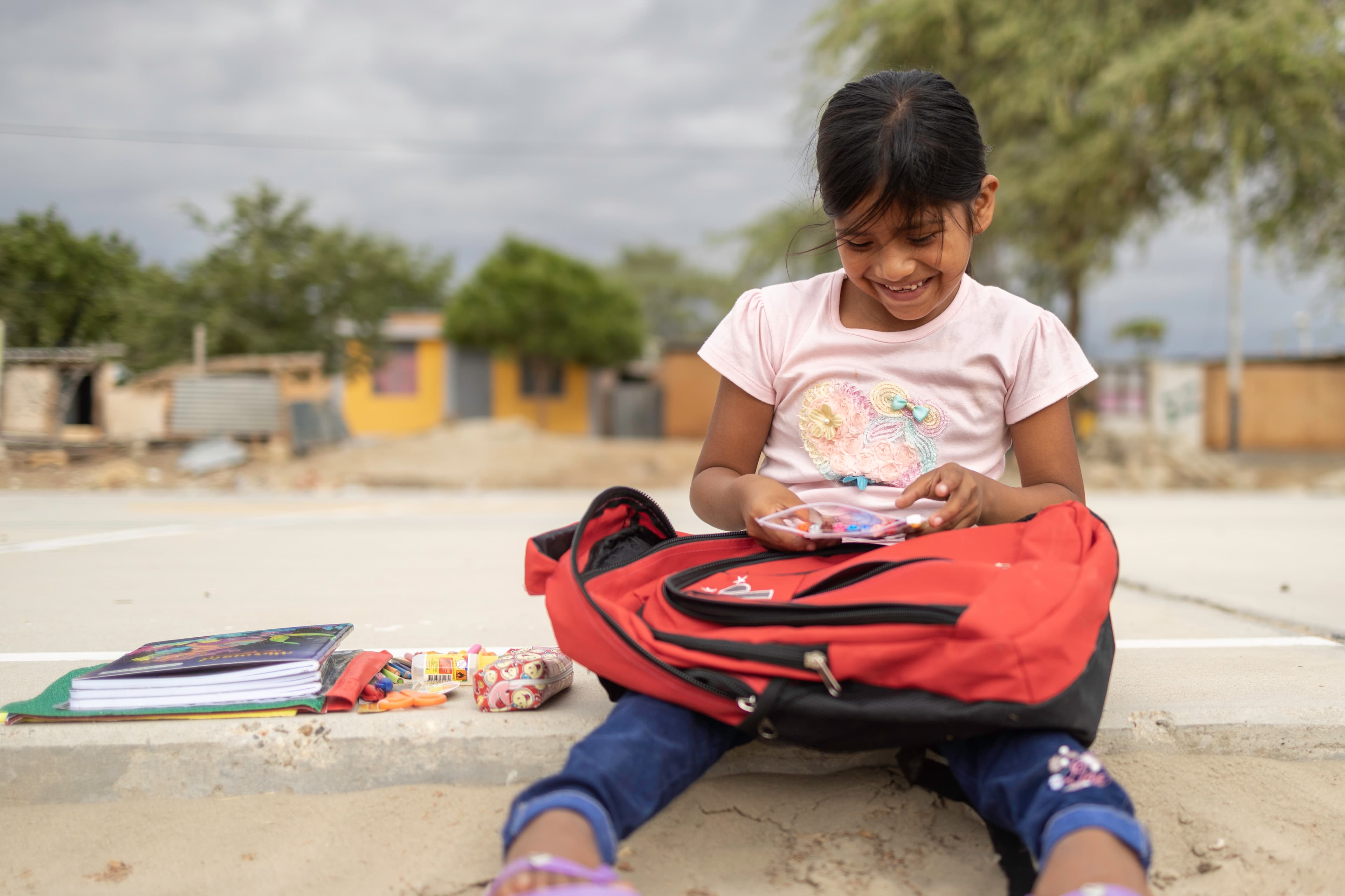 A young girl sits with a red backpack in her lap as she smiles.