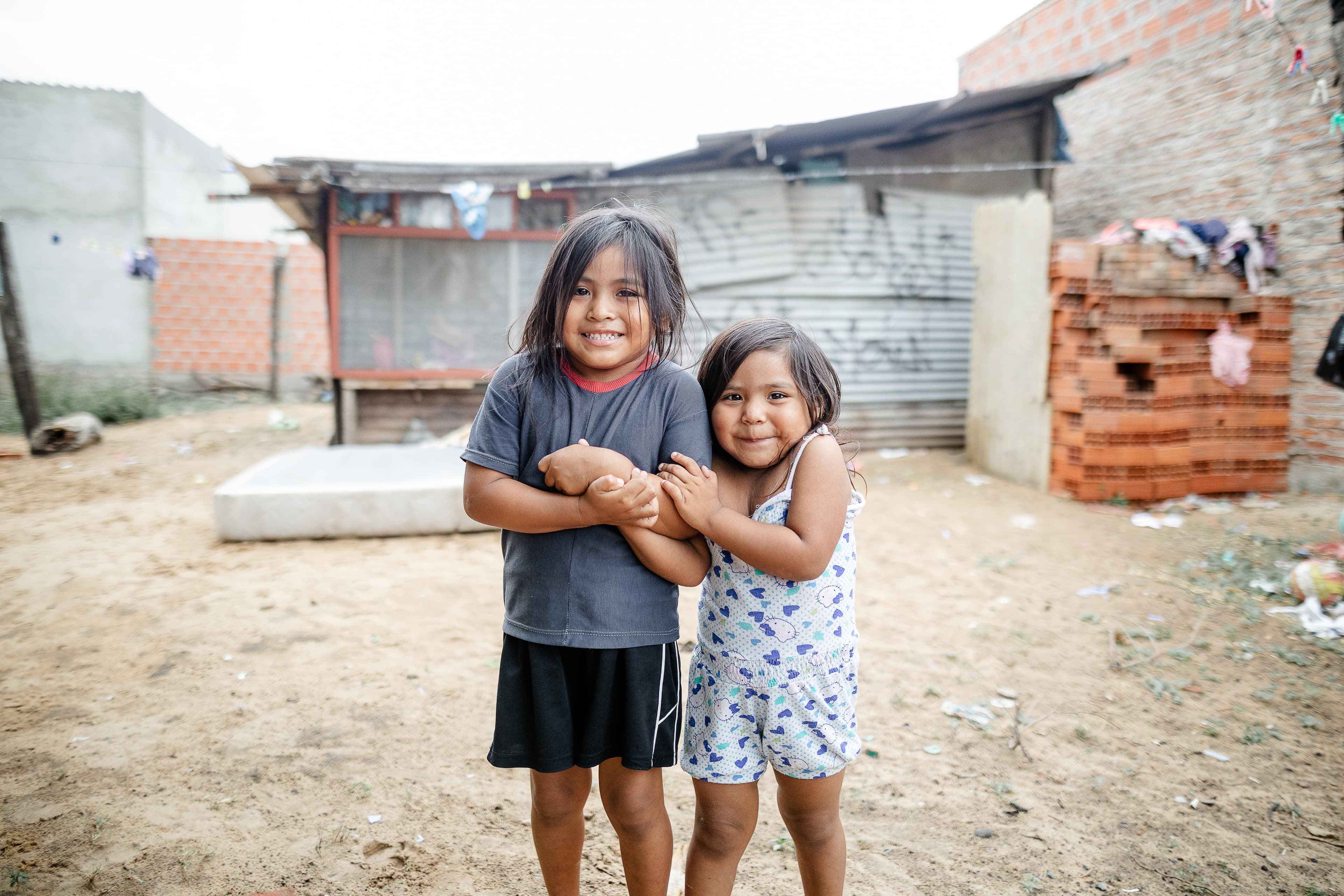 Two young girls embrace while standing in front of a tin home.