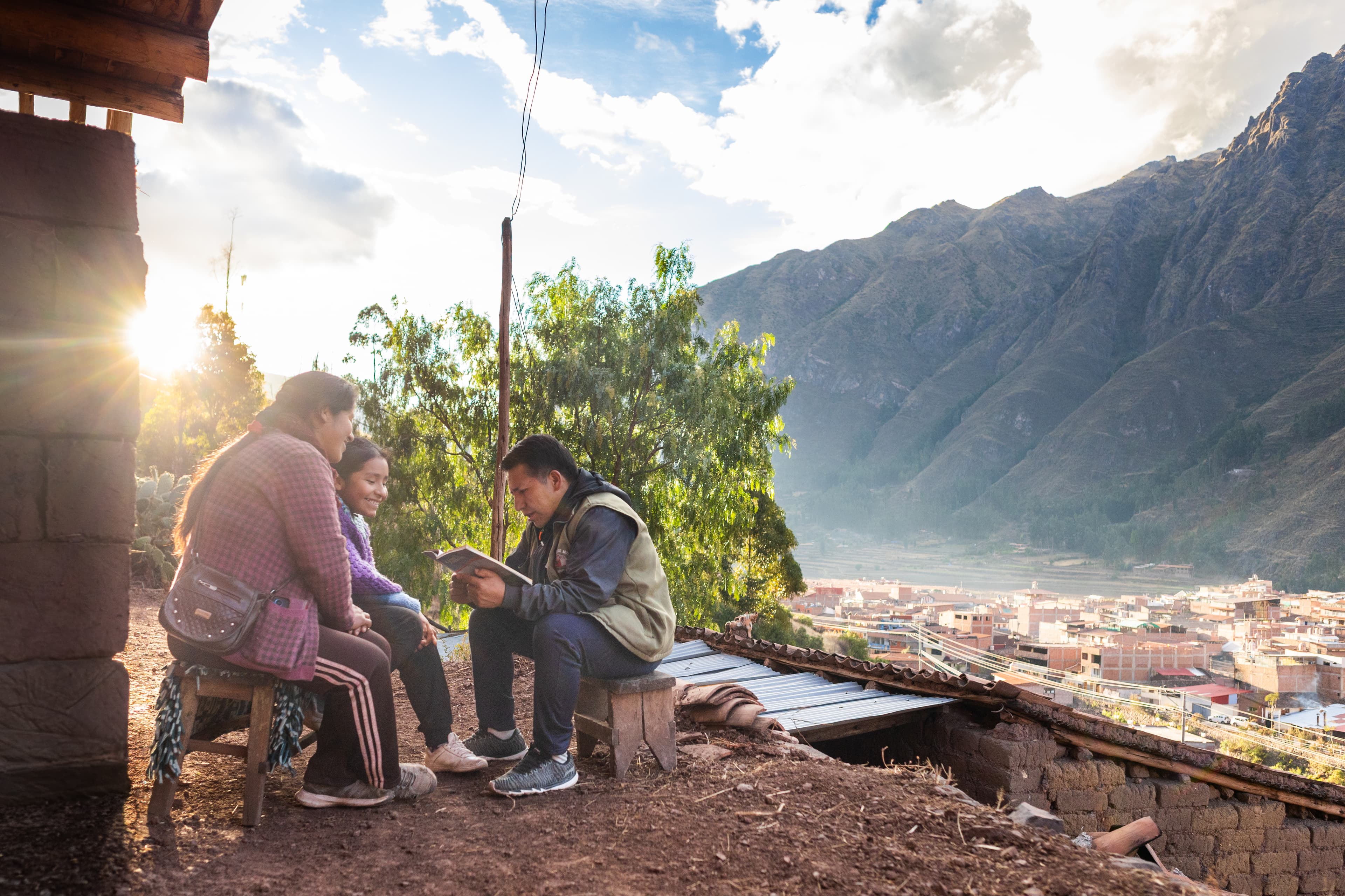 A man sits on a stool across from a woman and her daughter reading the Bible he is holding and behind them the sun shines through trees and to the right of them is a mountain over a valley of buildings.