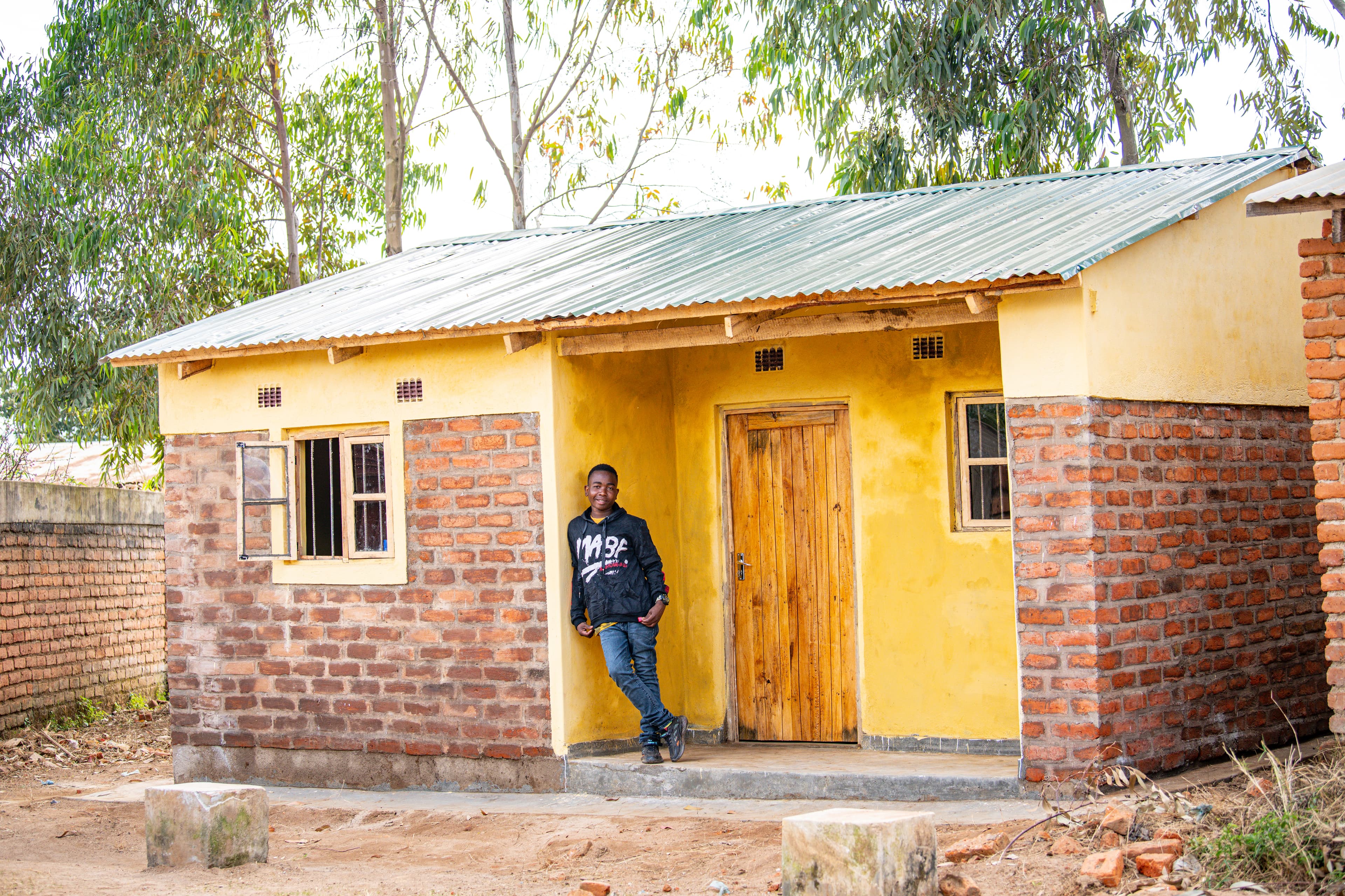 An African boy stands outside of a bright yellow home while smiling.