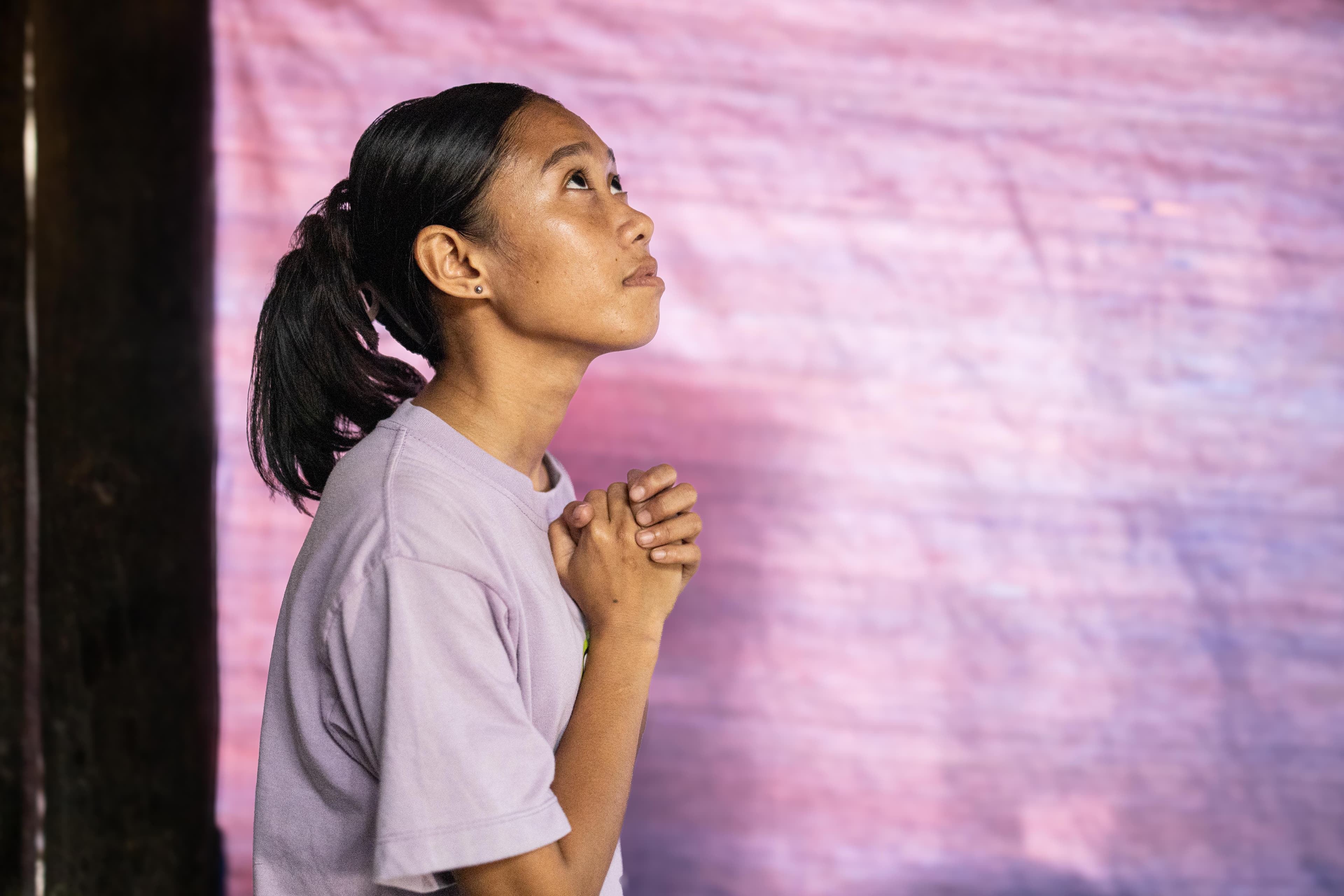 An adolescent girl wearing a purple shirt folds her hands in prayer and looks up. There is a purple background behind her.