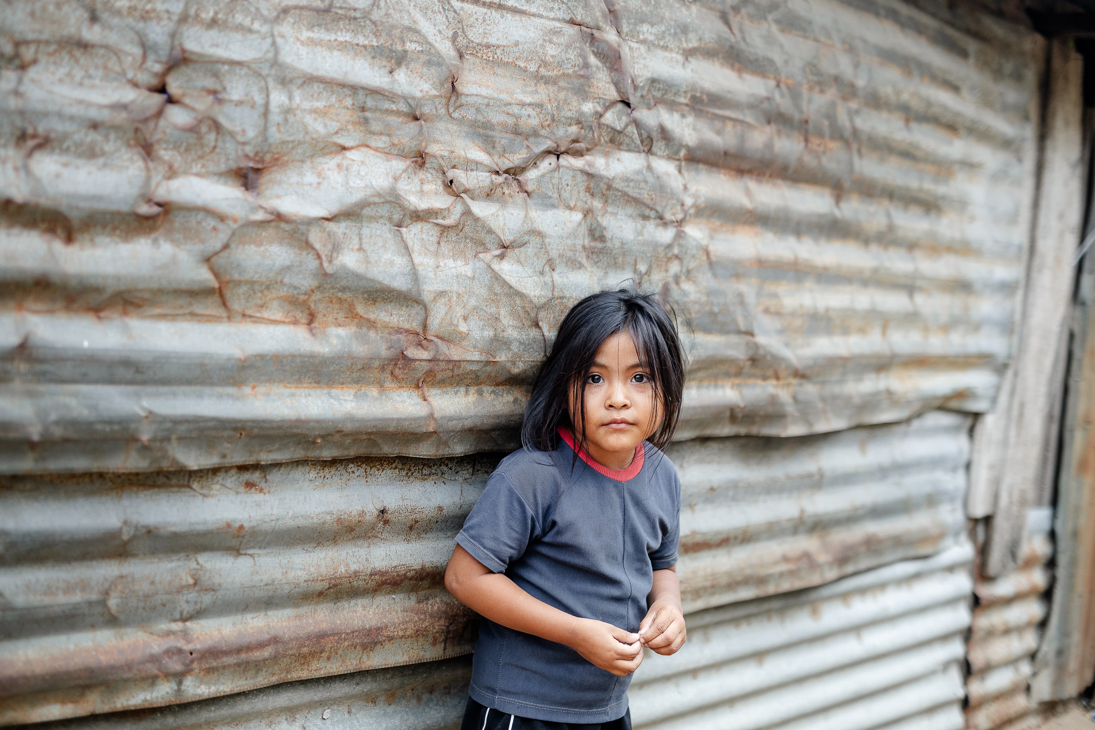 A young girl stands against a metal wall looking at the camera.