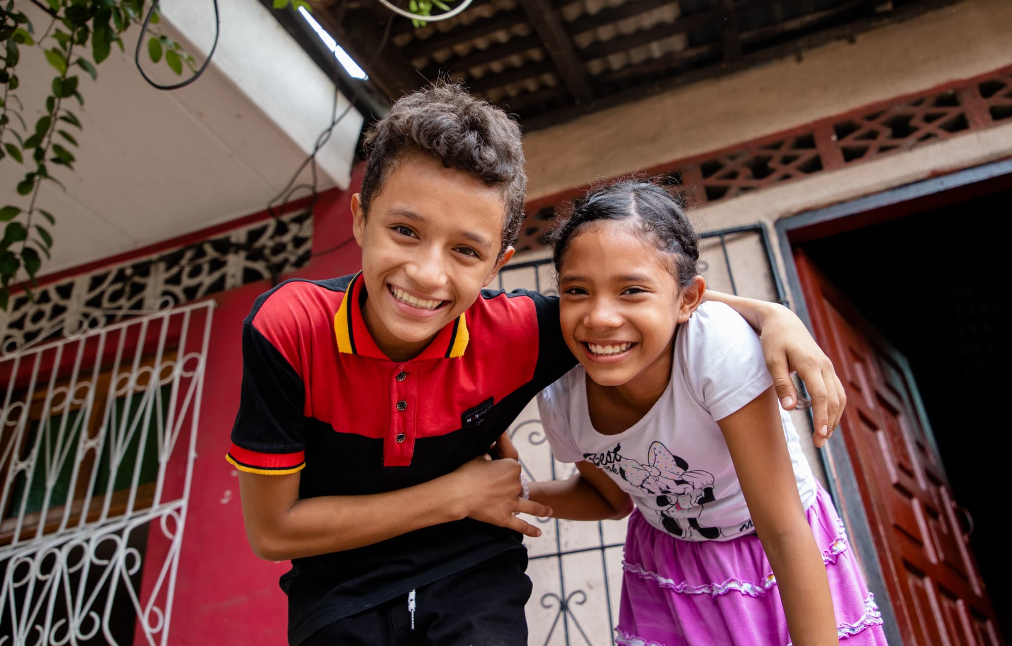 Teenaged boy—in black and red—and girl—in white and pink—lean forward and smile with arms wrapped around each other.