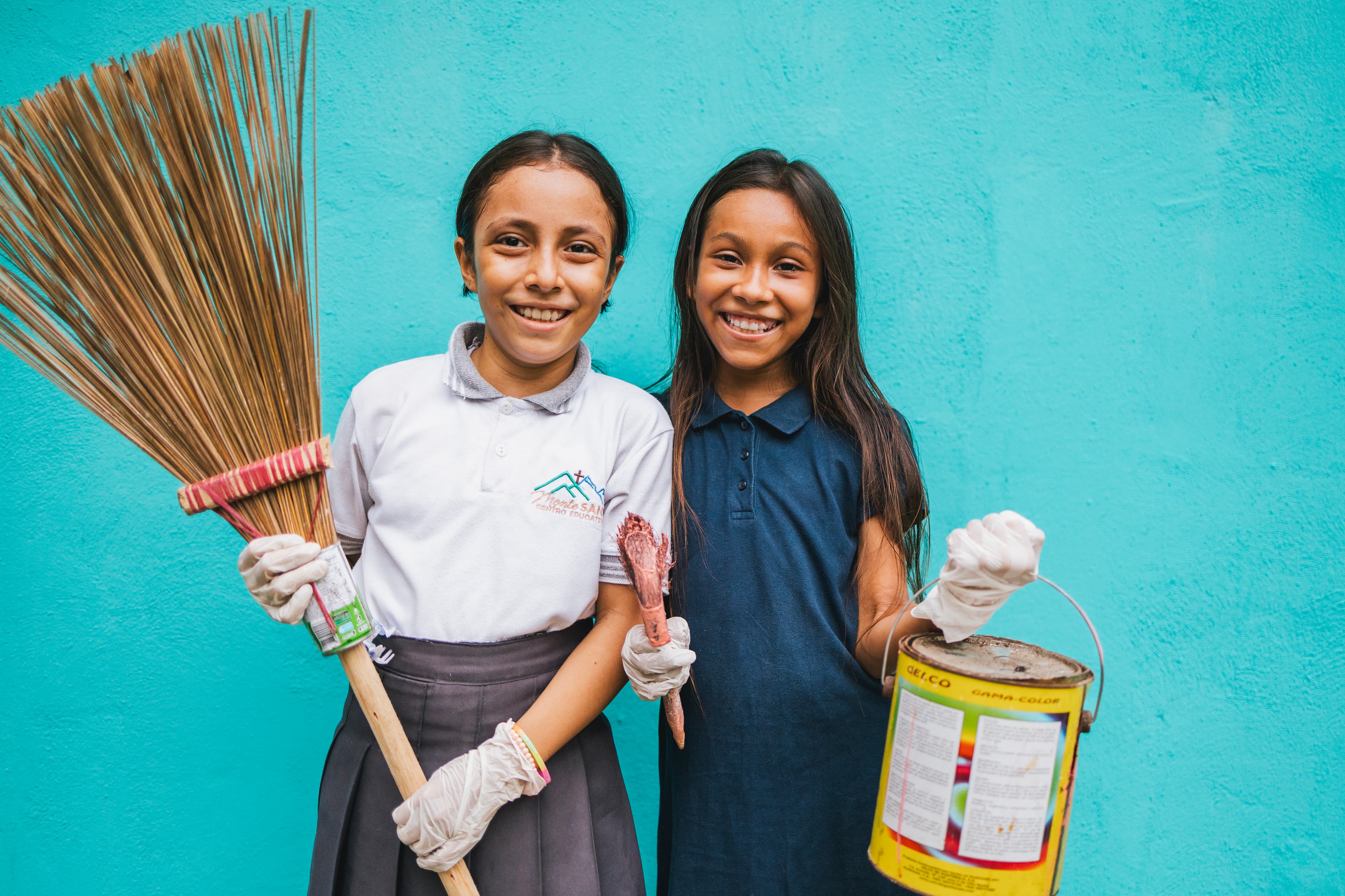 Two girls smile while holding a broom, paintbrush and a paint bucket again a blue green wall.