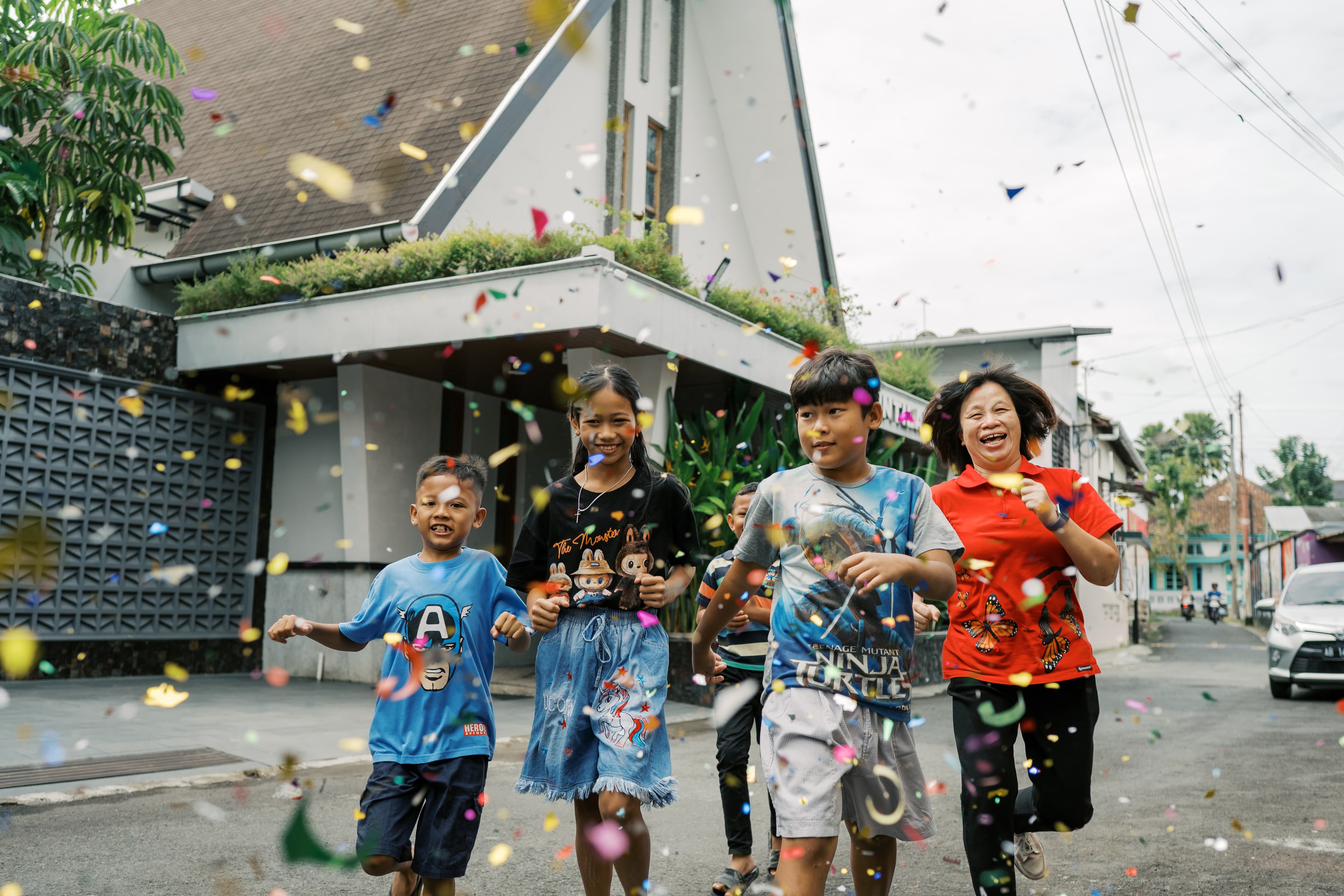 Four children and a woman walk down the street in front of a church, with colorful confetti falling around them.
