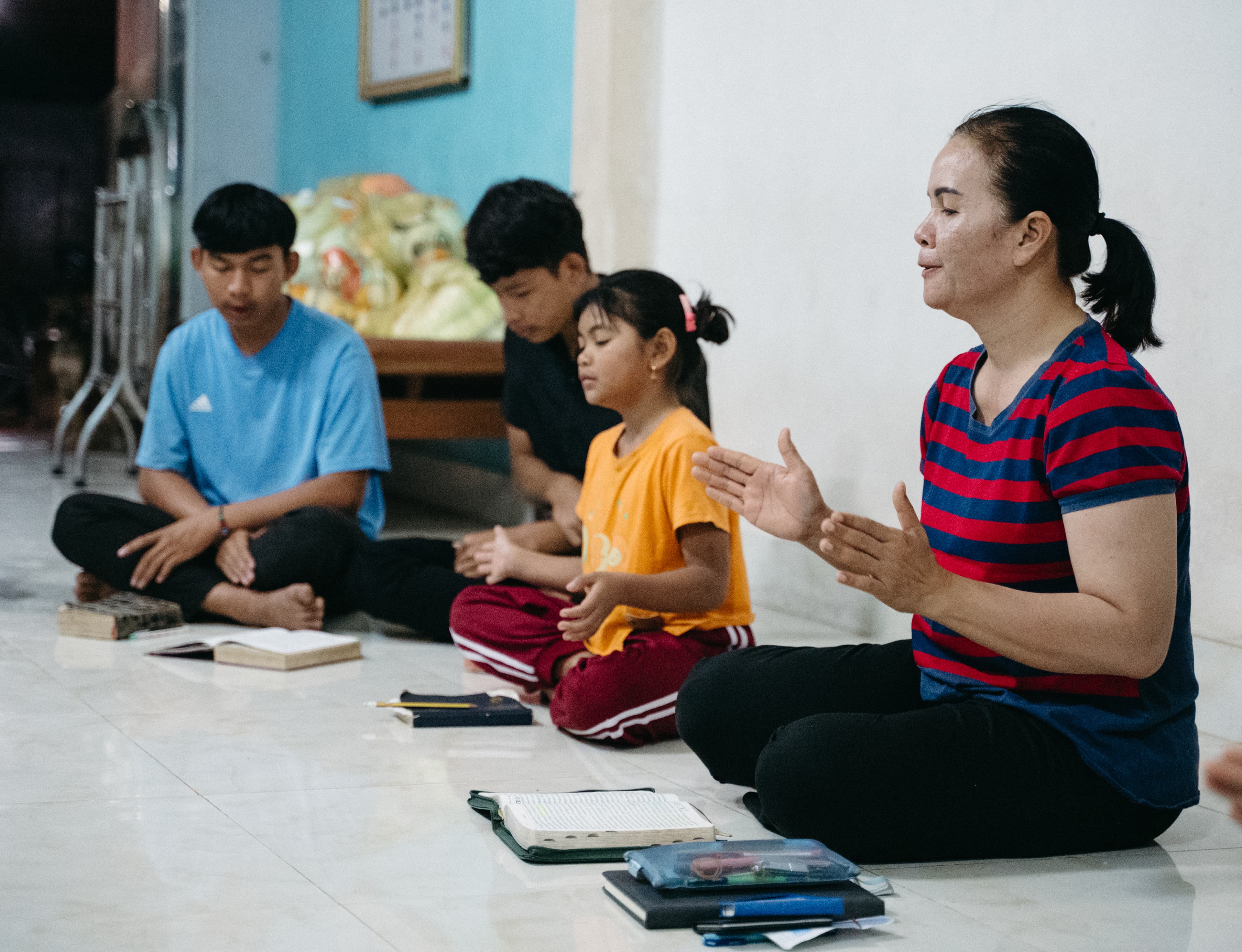 A woman sits cross-legged on the floor wearing a red and blue striped shirt, singing with a group of youth and students in a church setting.
