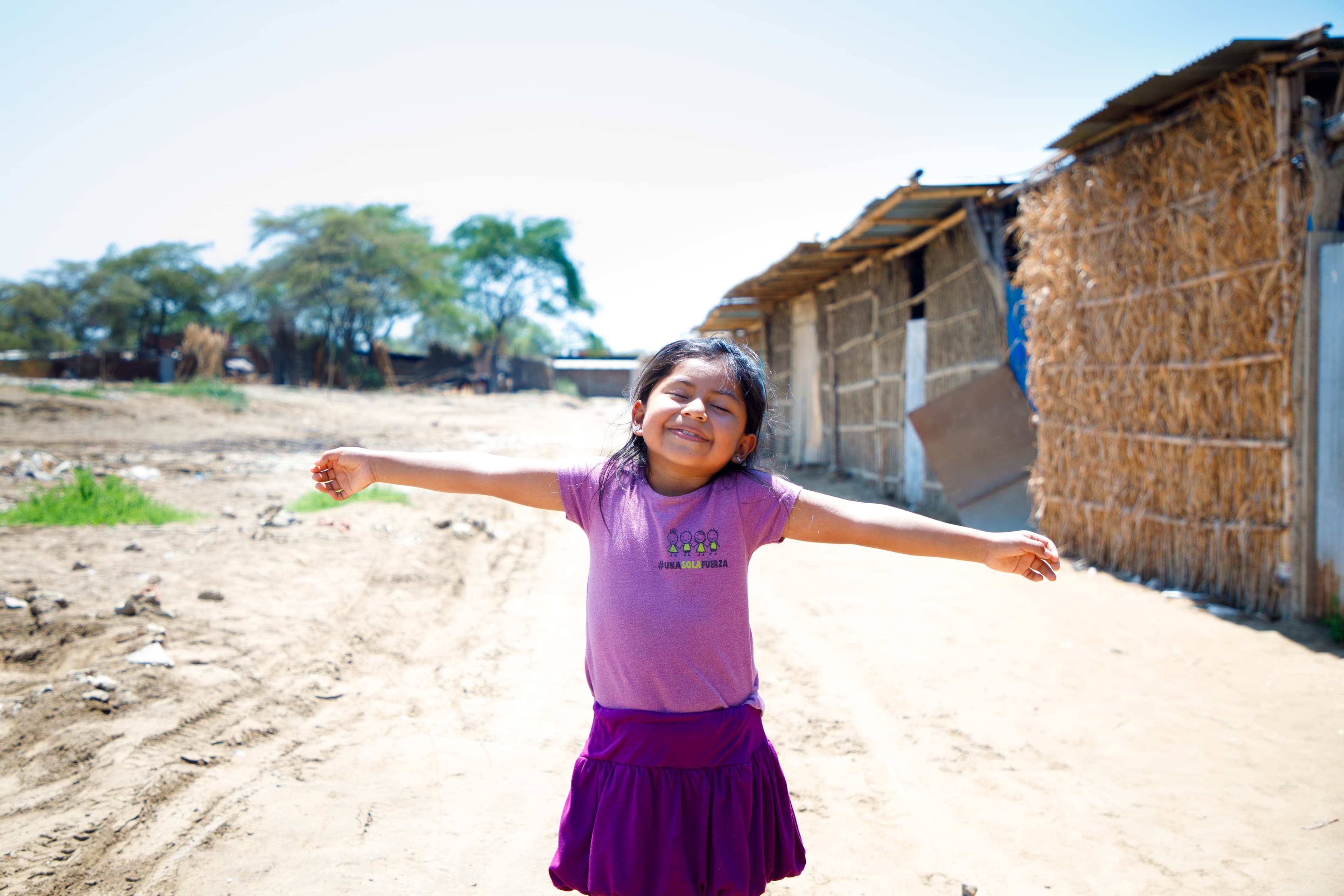 A young girl wearing purple stands with her arms outstretched while standing in a dirt road.