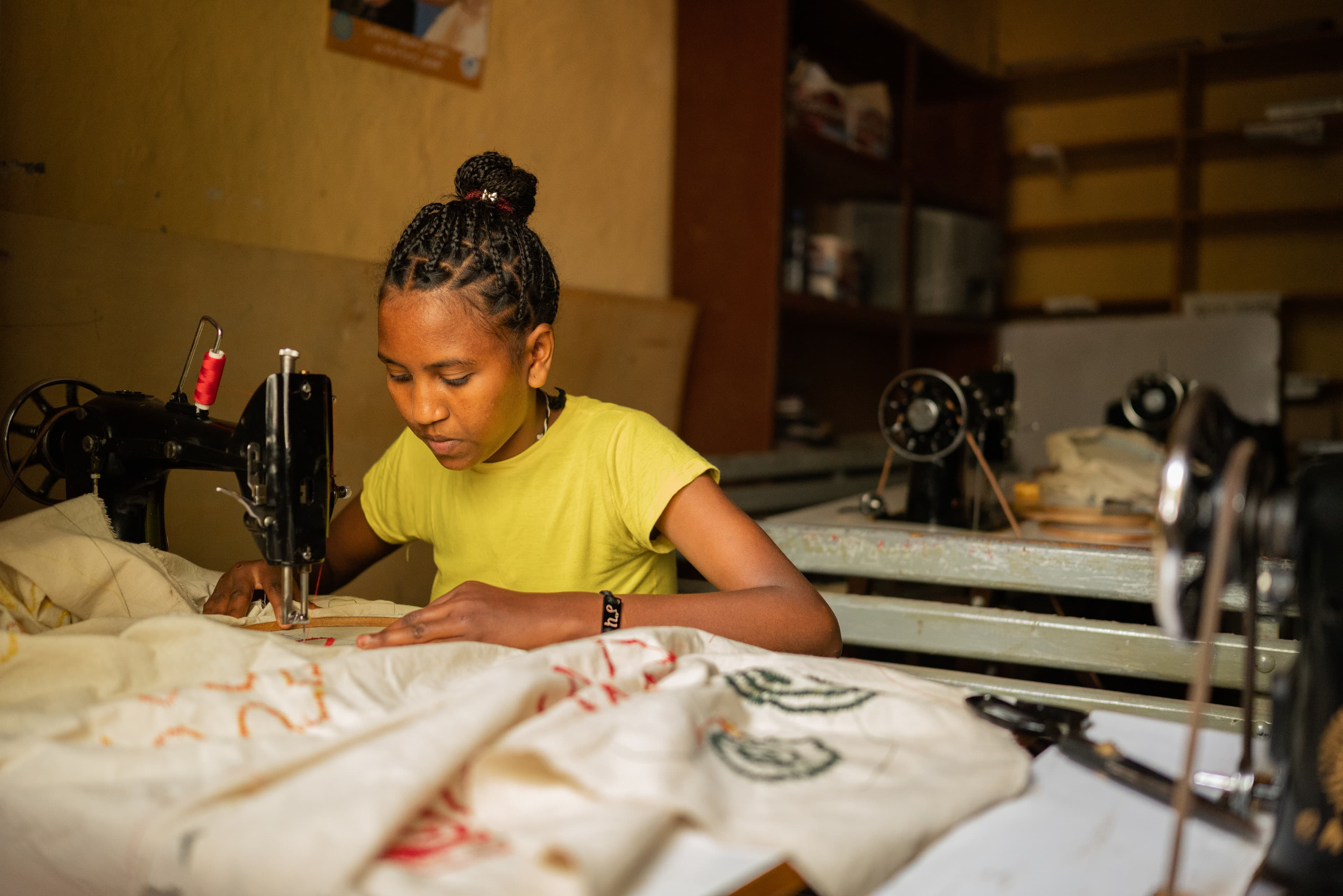 Teenaged girl wearing a yellow shirt sits at a sewing machine with fabric.