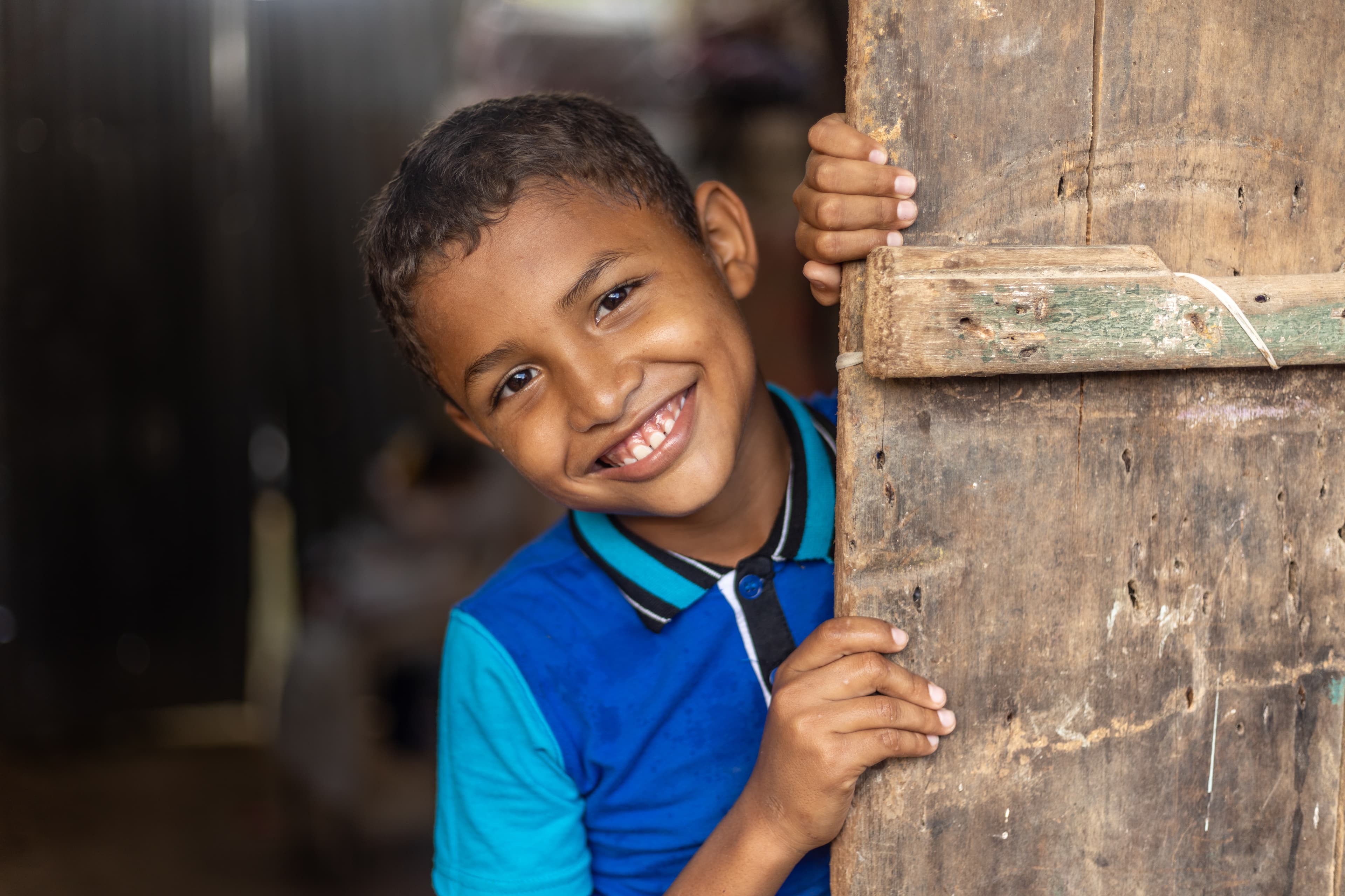 A boy wearing a blue shirt smiles as he looks out from behind a wooden wall