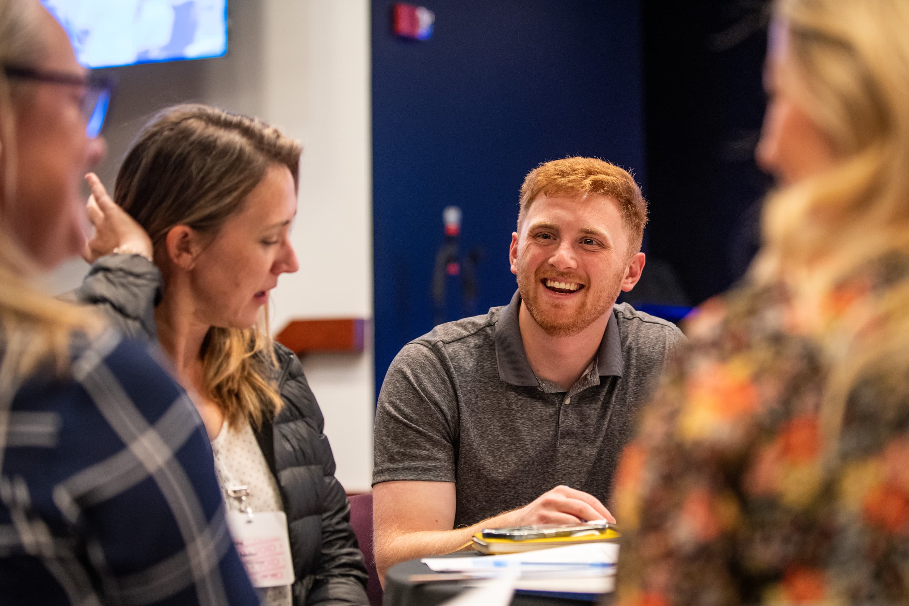 A young man sits at a table with other colleagues while smiling.