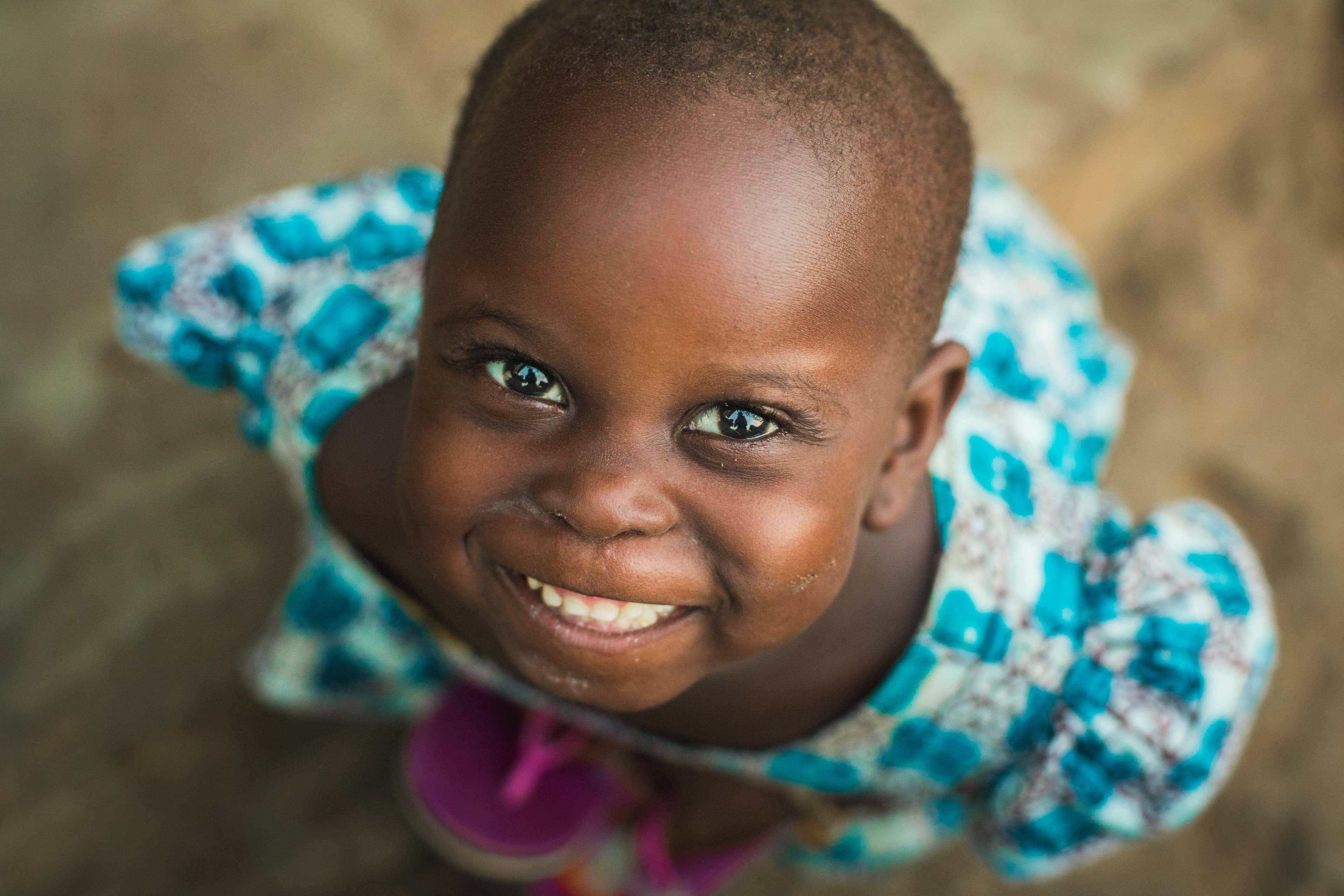 A young African girl wearing a blue and white dress looks up at the camera and smiles.