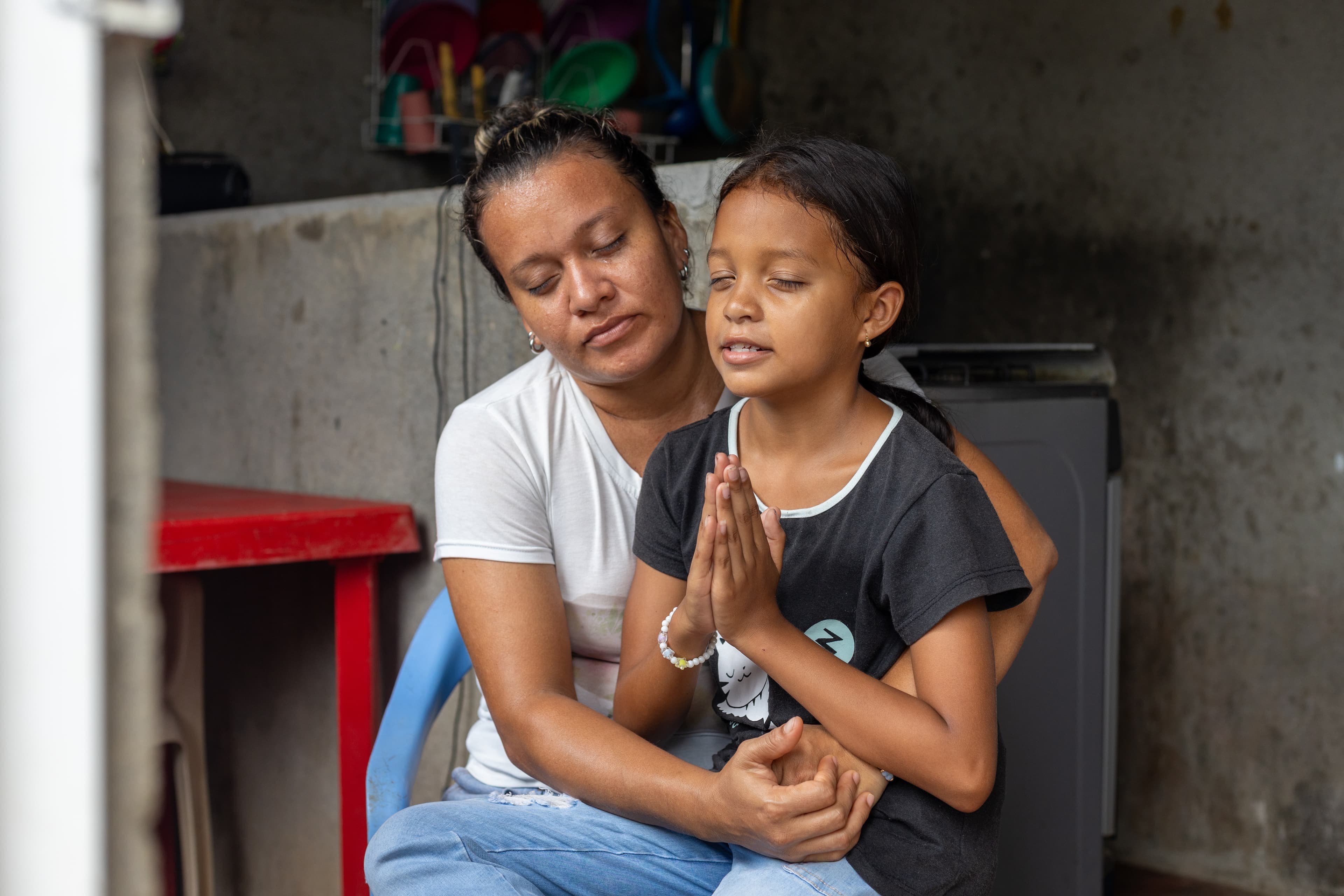 A young girl sits on the lap of an older woman as they both pray together.