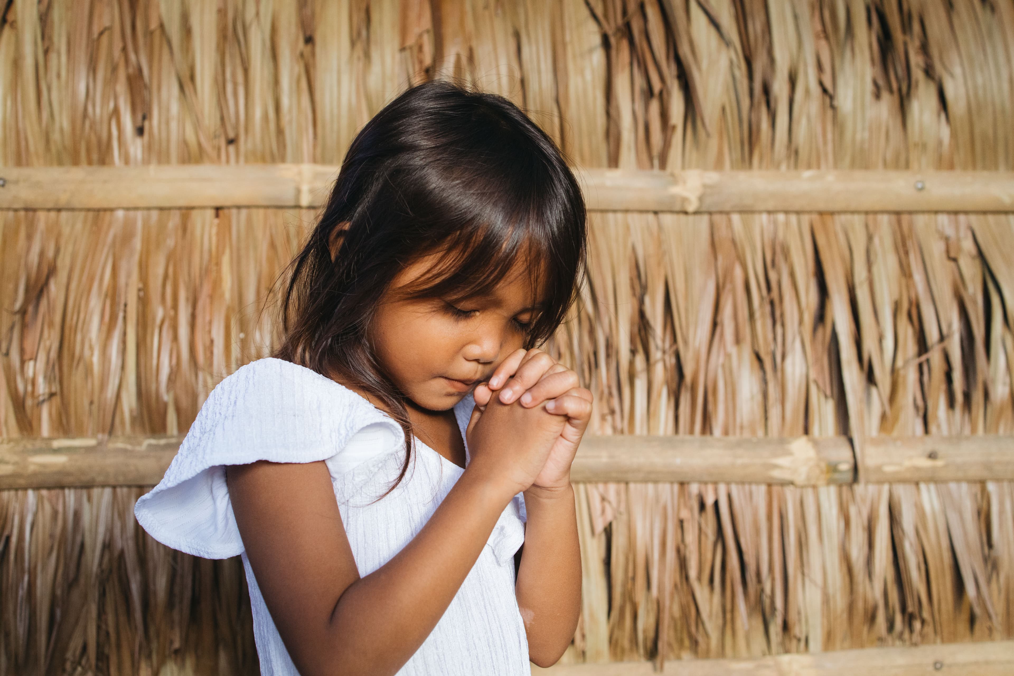 A girl in a light blue dress prays in front of a straw background.