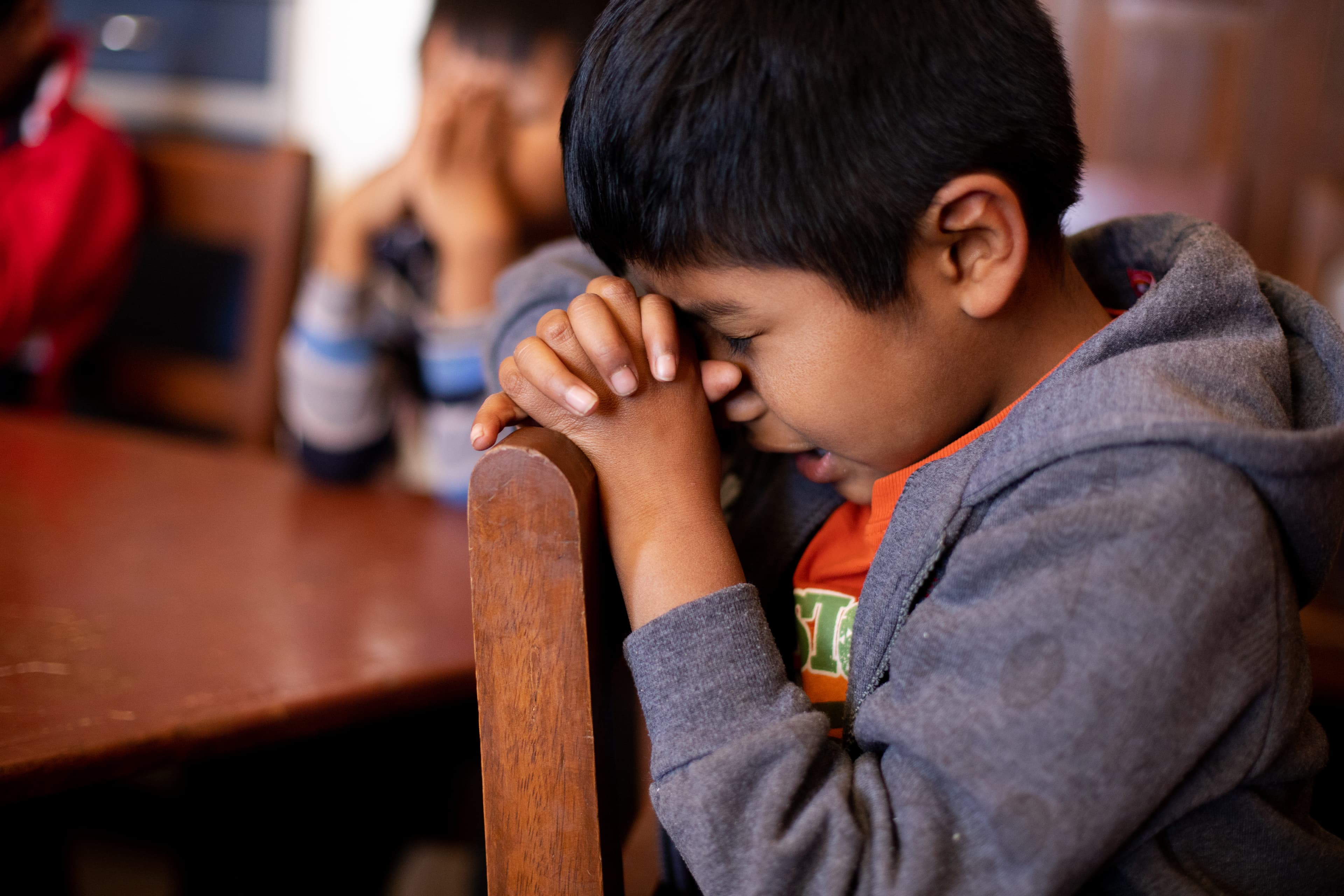 A young boy wearing a gray jacket sits and prays on a chair with his eyes closed and hands clasped against his face.