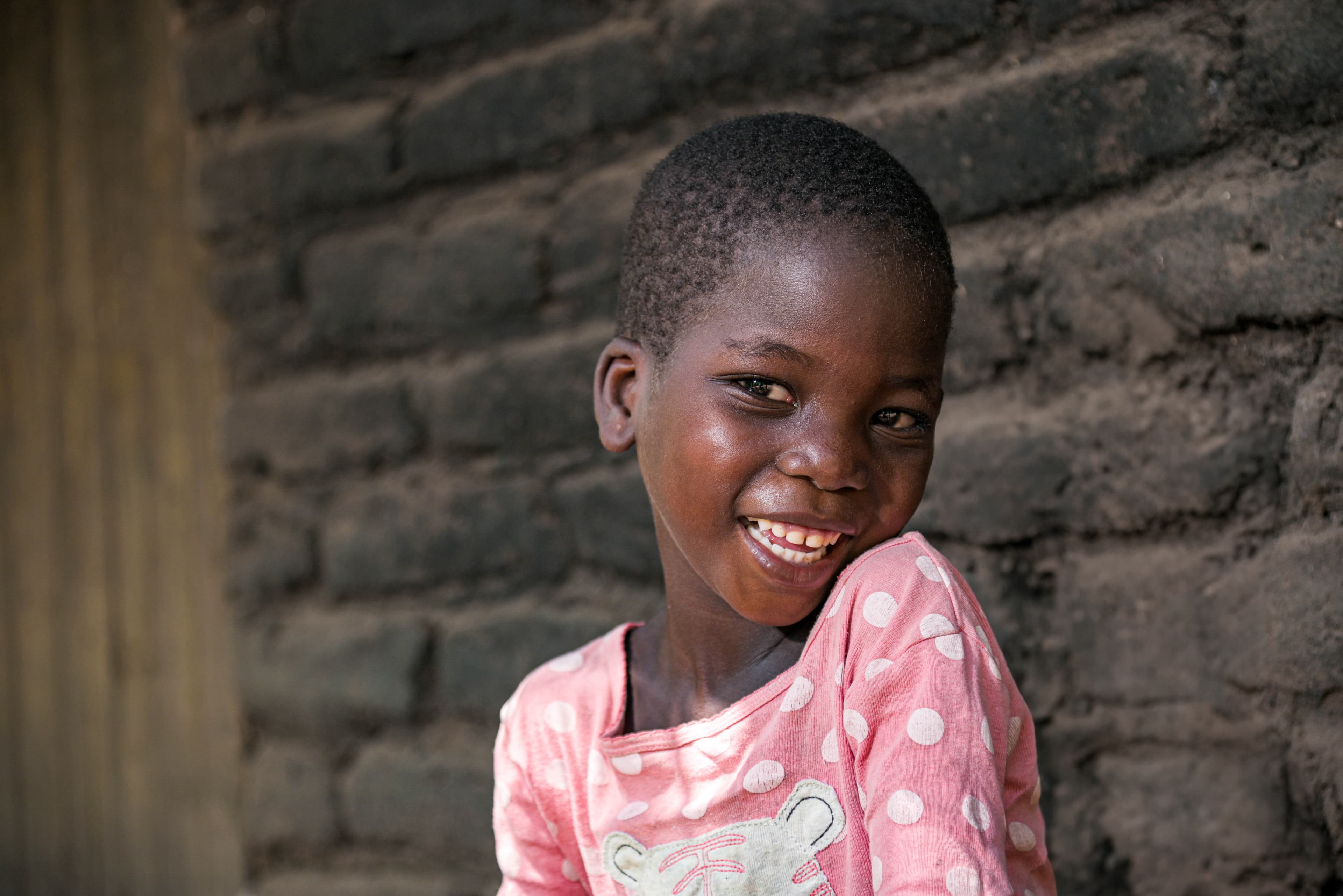 A young girl is sitting against a wall smiling at the camera.