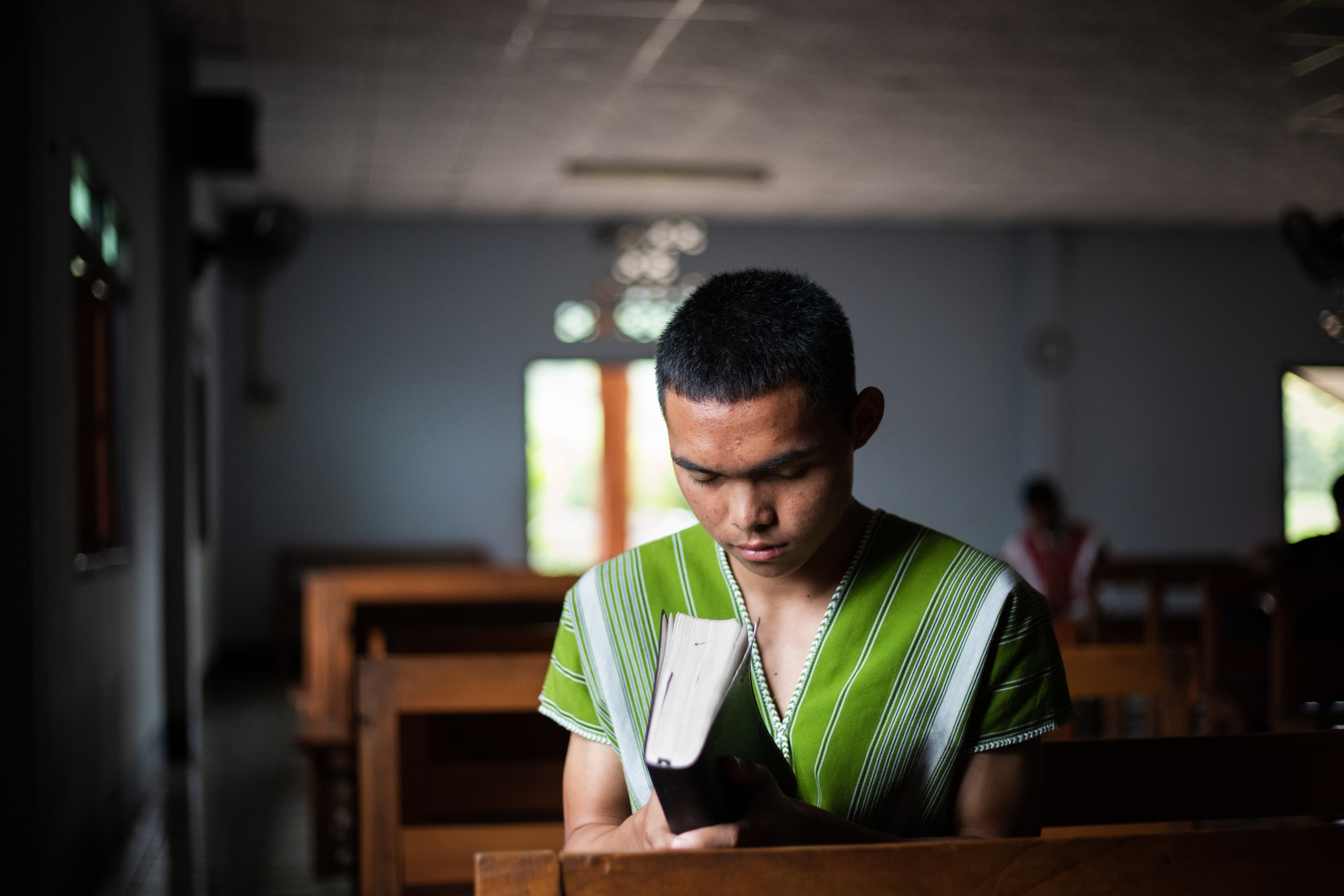 A young man sits at a desk praying as he holds a bible.