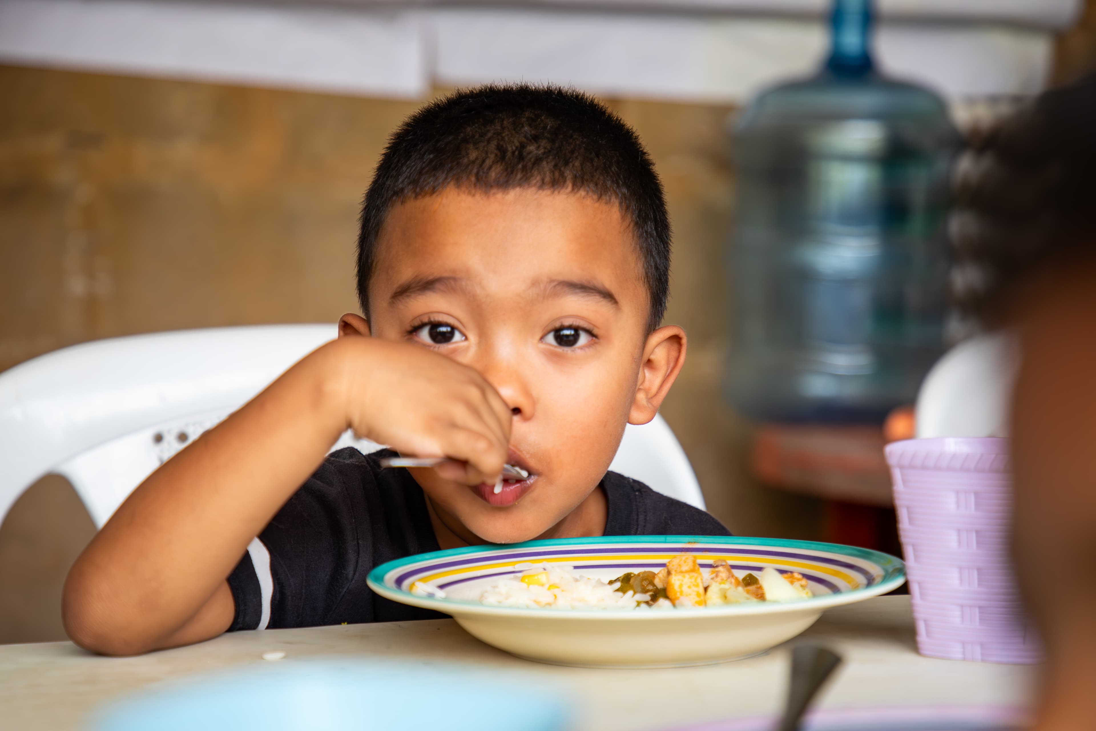 A young boy sits behind a bowl of food while using a spoon to take a bite.