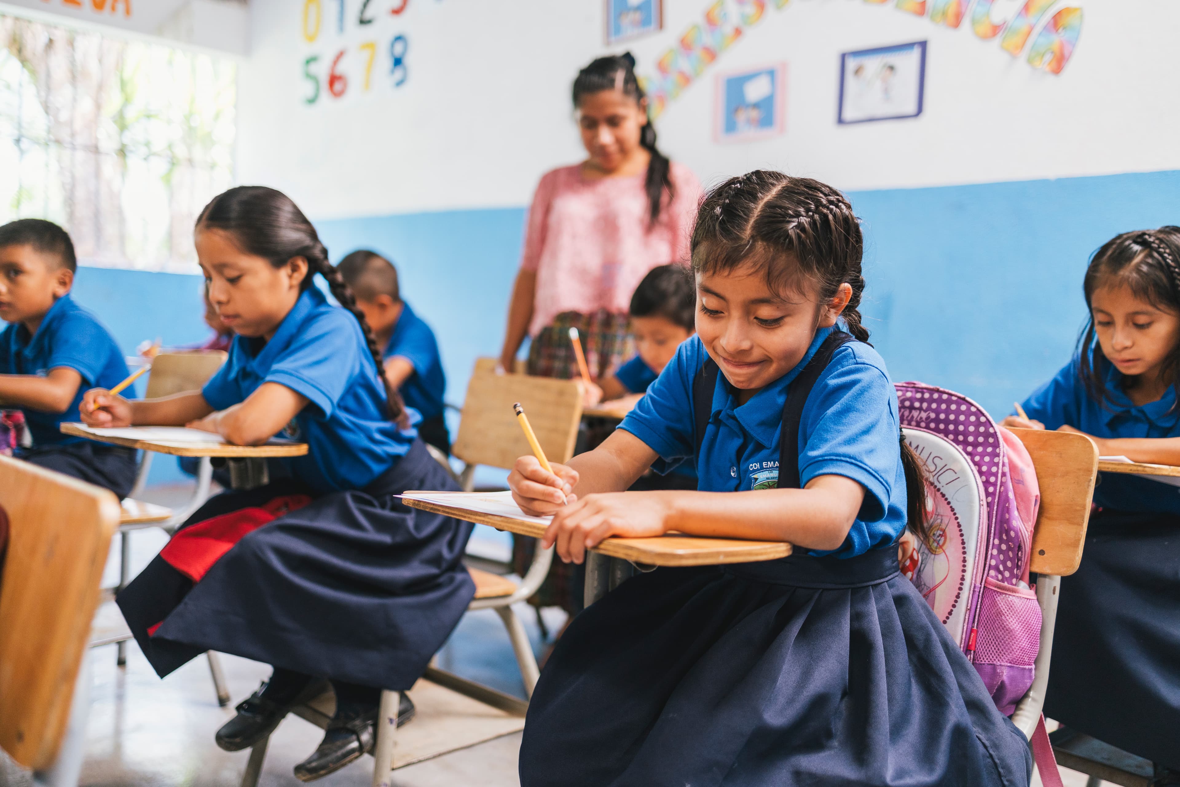 Young girls wearing school uniforms sit at desks while writing and smiling.