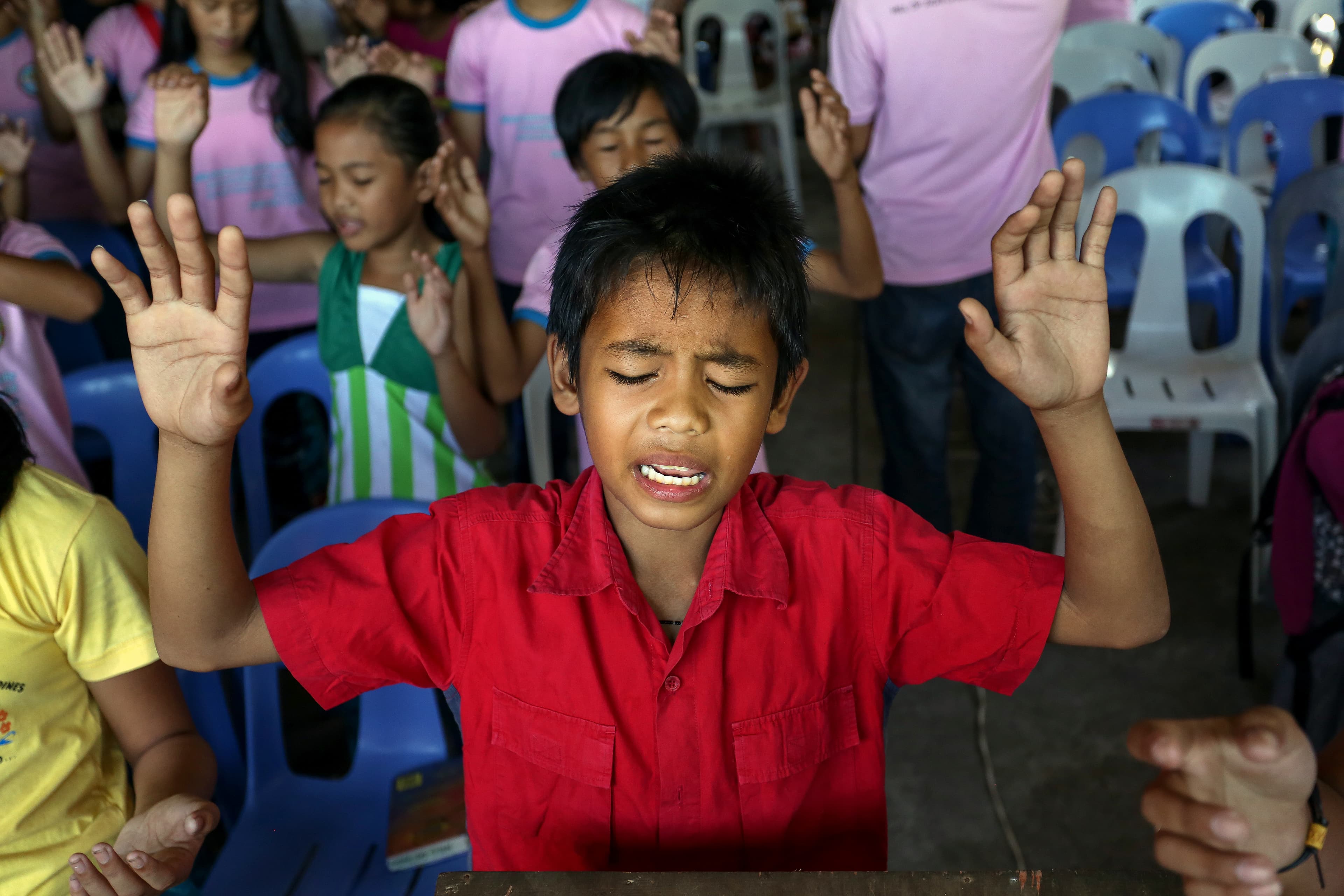 Young boy wearing a red shirt lifts his hands in worship with other children during a church service.