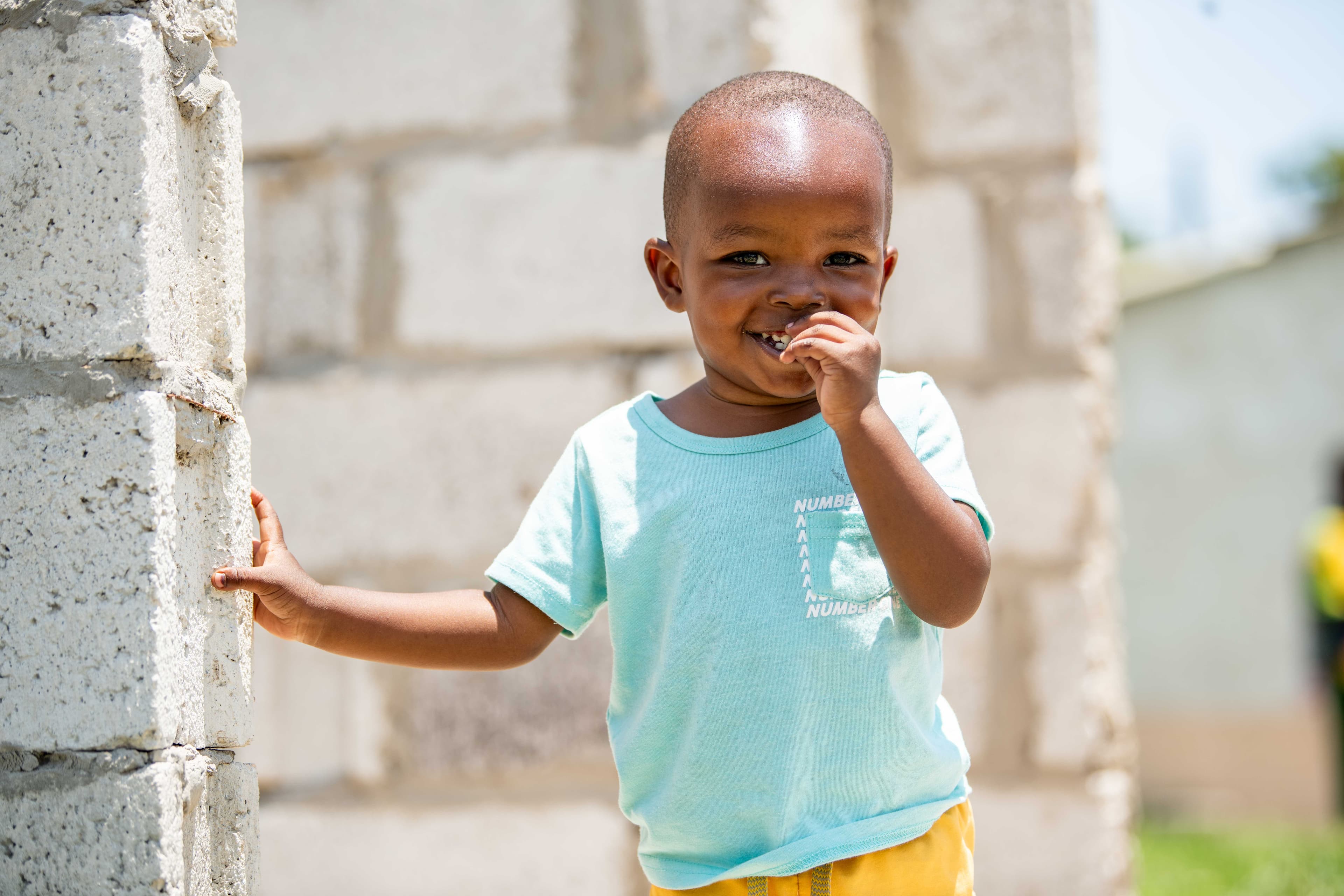 A young boy wearing a blue shirt smiles for the camera while holding onto a block wall.