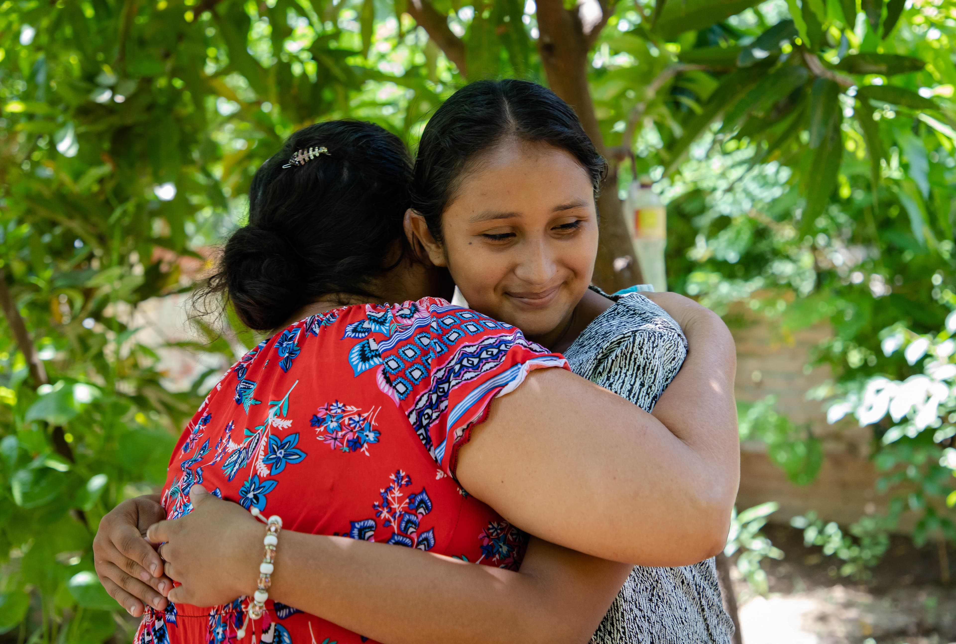 A teenaged girl hugs an older woman wearing a red patterned shirt. Trees are in the background.