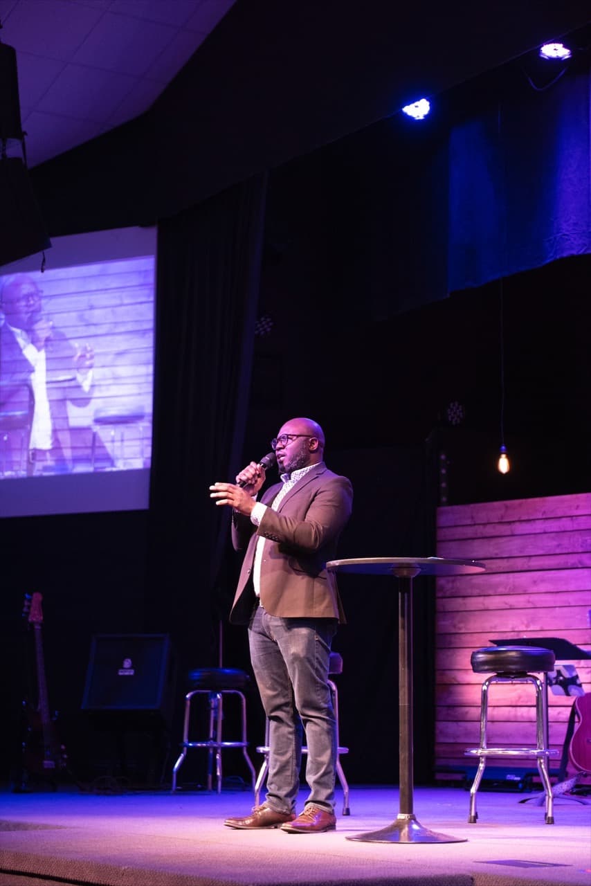 A man wears a brown suit jacket and stands on a stage holding a microphone with a purple background and screen behind him.