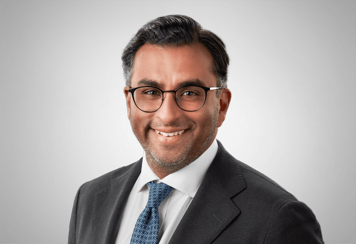 A man in a black suit and blue tie smiles in front of a black background.